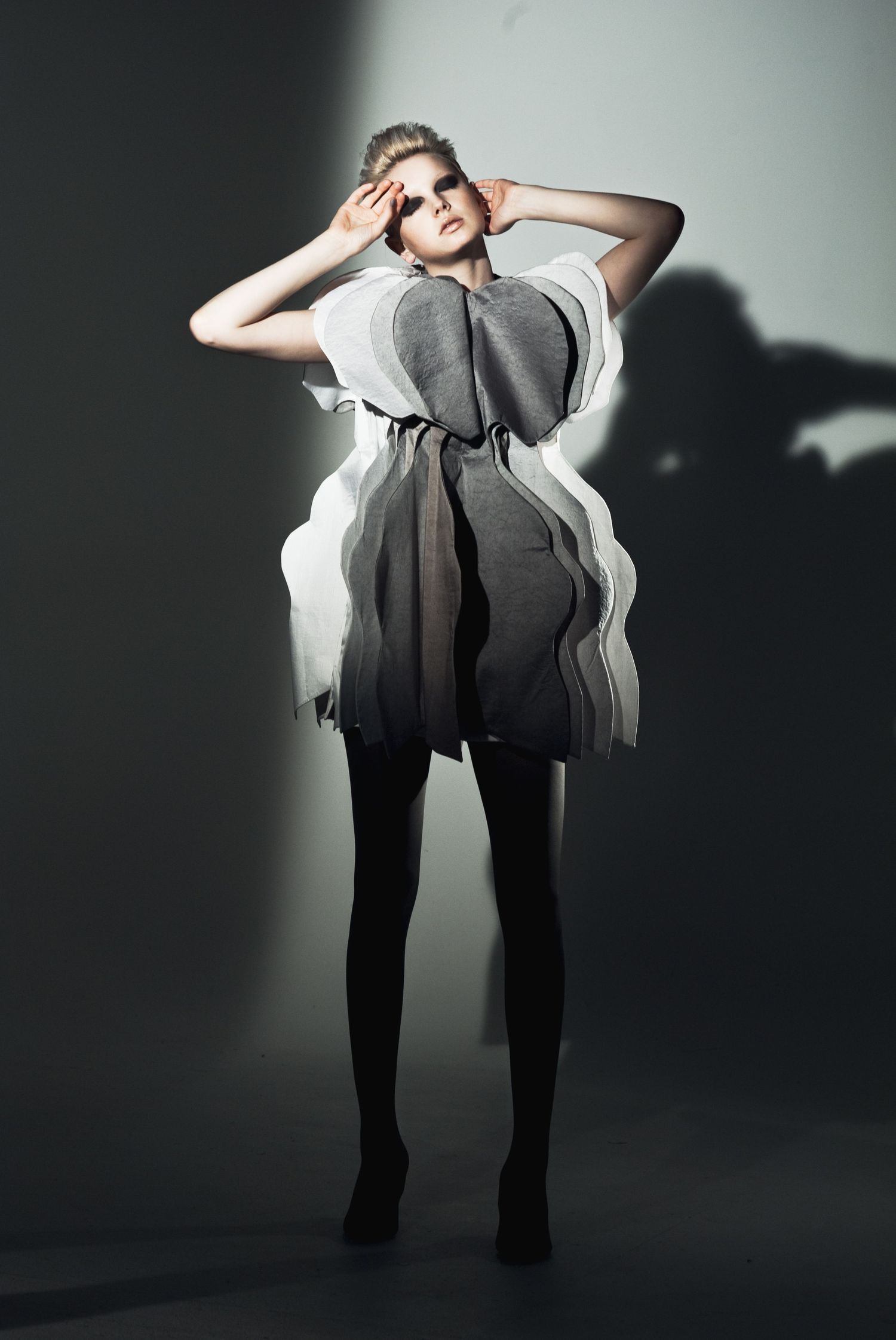 Model wearing avant-garde sculptural gray and white layered dress with black tights in dramatic studio lighting.