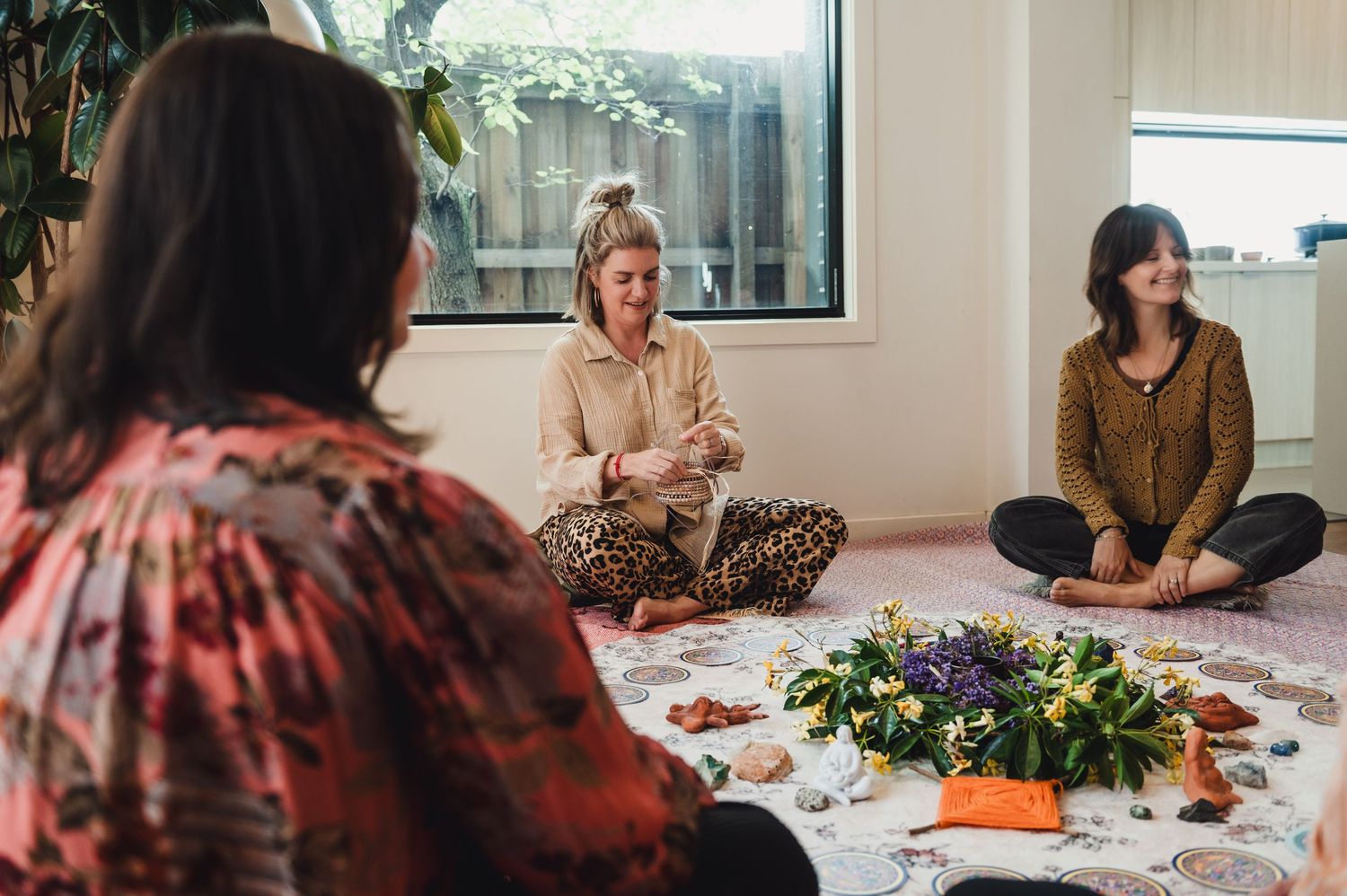 Three women sit cross-legged in a circle on a floral mat with flowers, engaged in a peaceful wellness group session indoors.