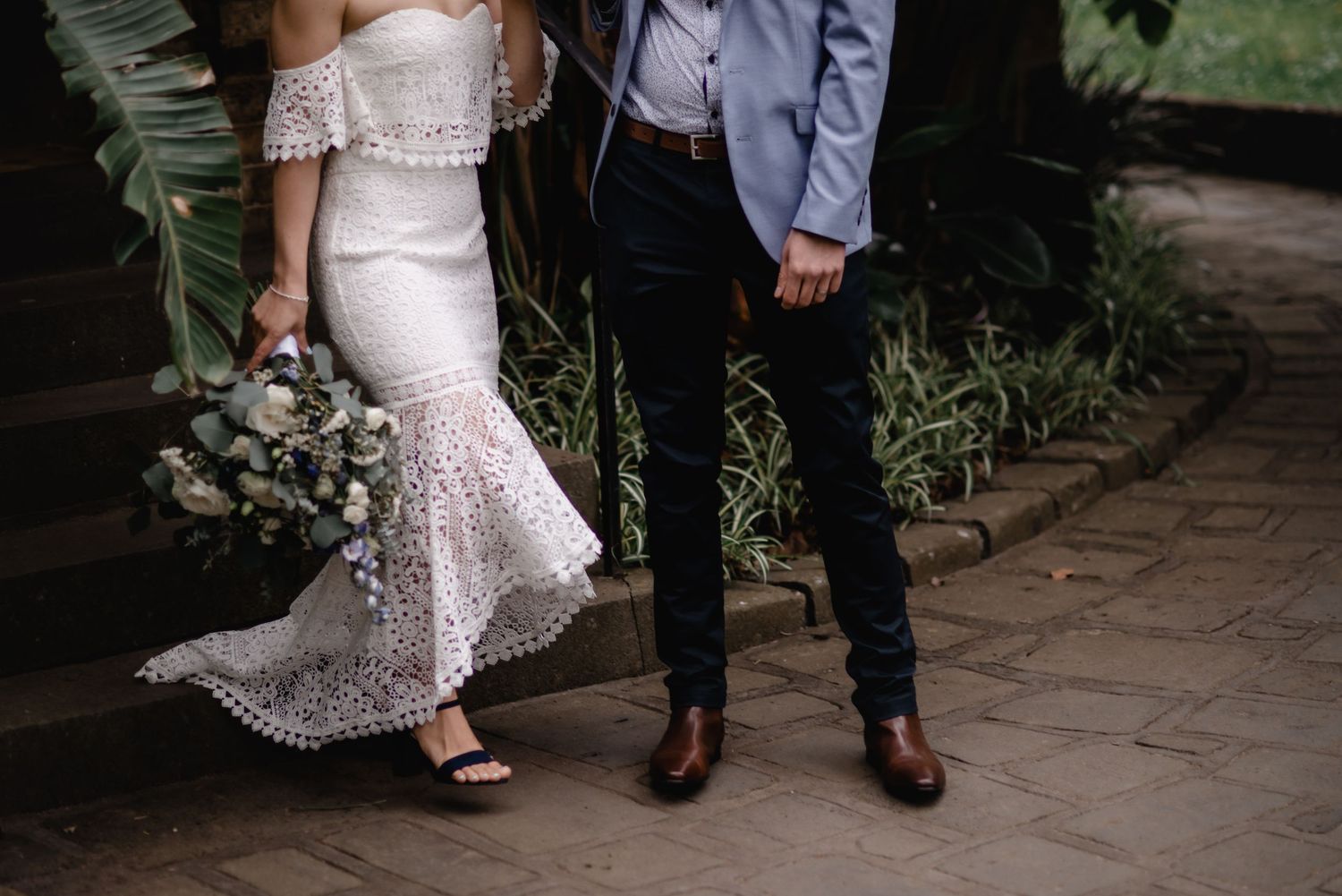 Detail shot of a lace wedding dress and suit with bouquet against stone pathway.