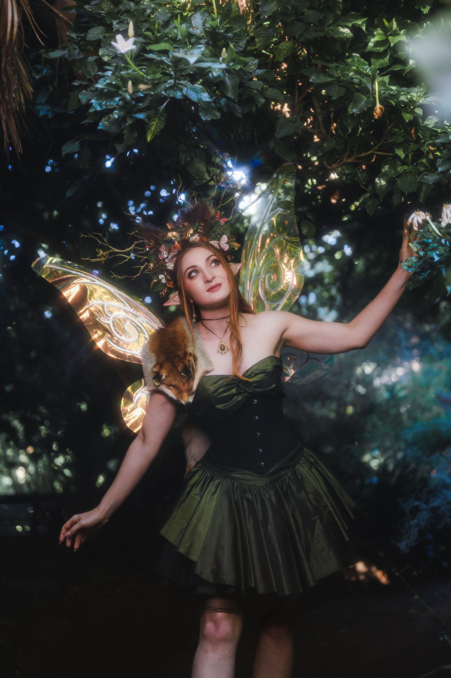 A woman in a green fairy costume with glowing wings and floral crown poses magically among lush trees at night.
