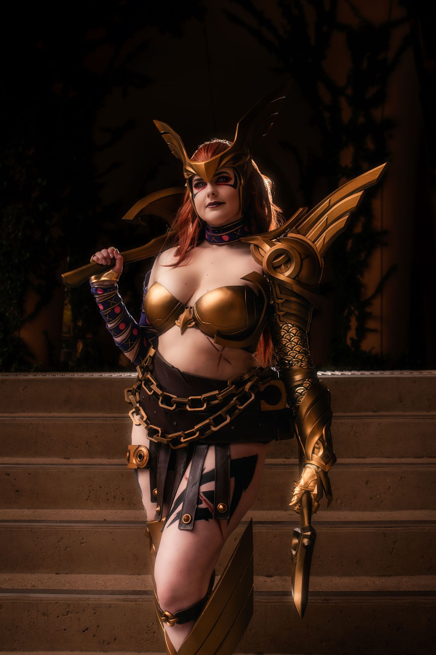 Woman cosplaying in golden armor costume with wings and chains, posing dramatically on stone steps.