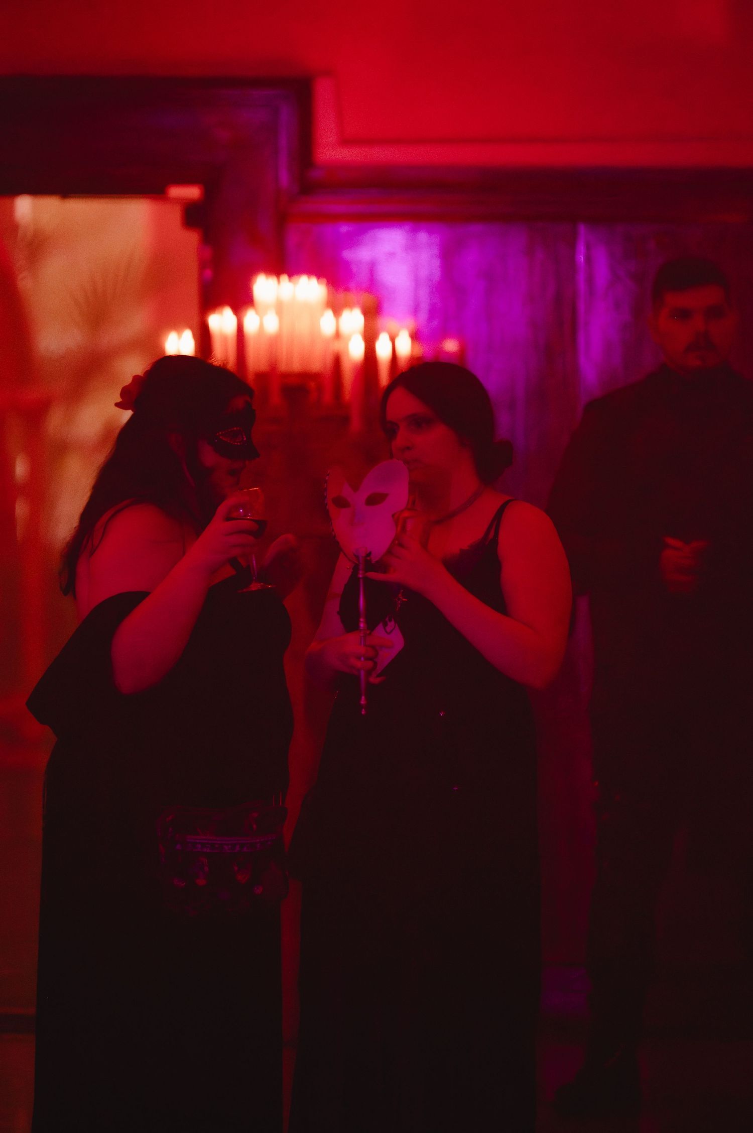 Two women in black dresses wearing masquerade masks hold drinks at a candlelit masquerade party bathed in red light.