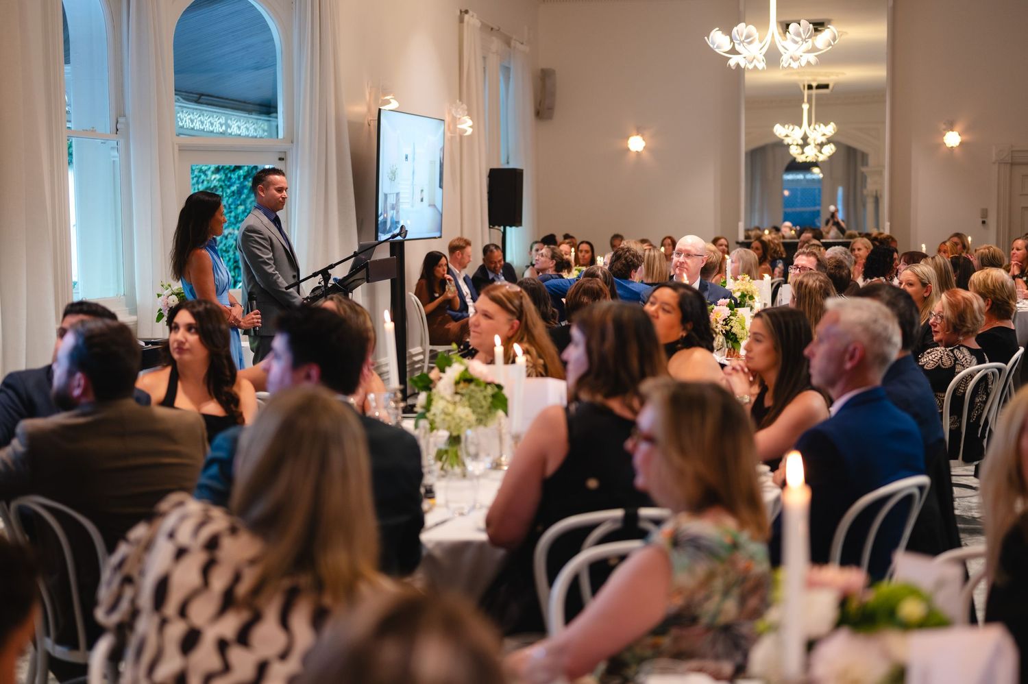 Wedding reception guests seated at elegant round tables in a bright banquet hall with chandeliers overhead.