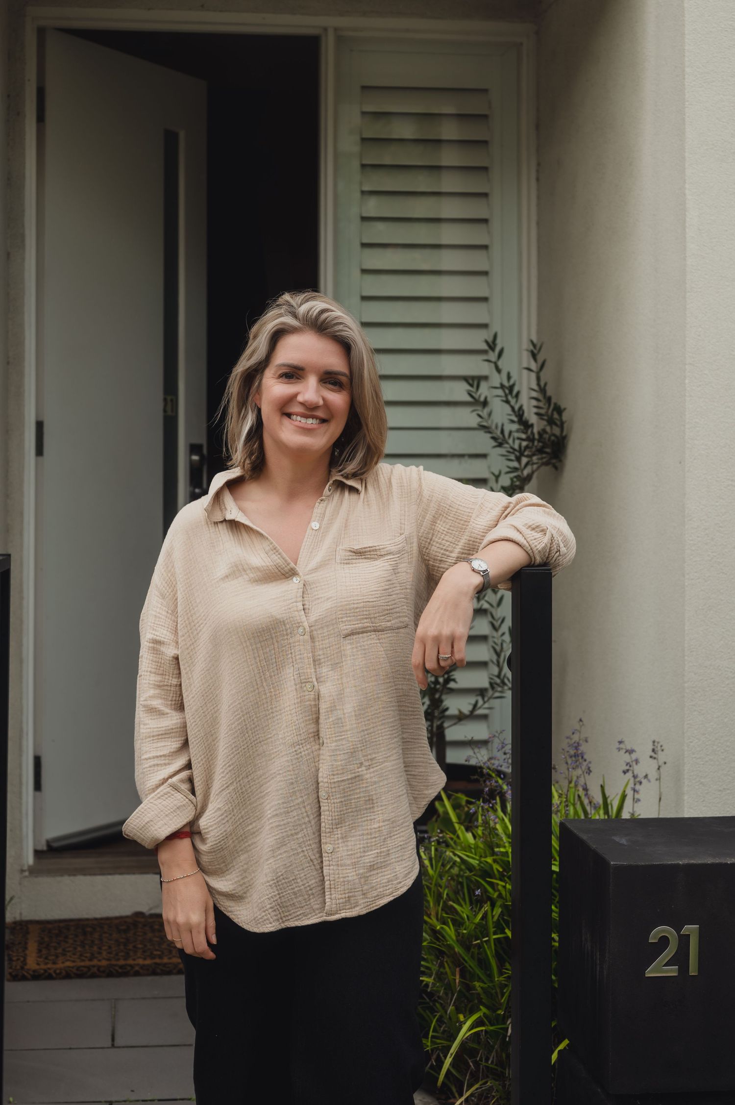 Smiling woman in beige linen shirt standing by mailbox numbered 21 outside a modern home entrance.