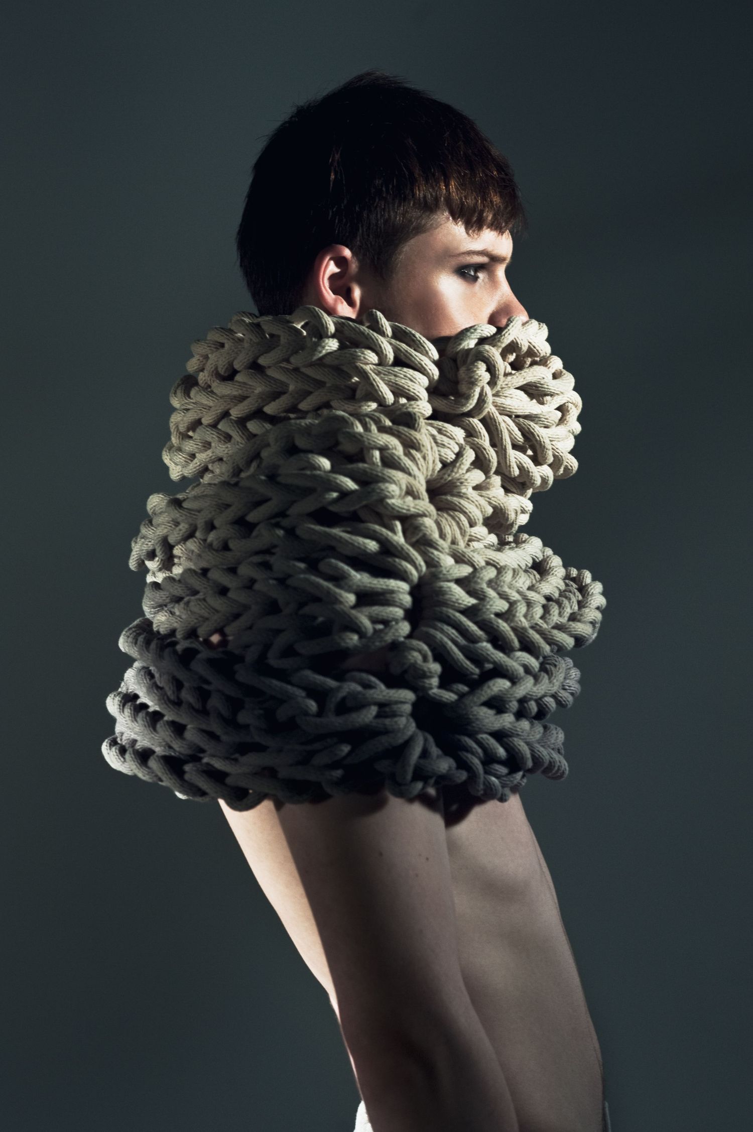 Person wearing an oversized chunky knit wrap in olive green, styled as avant-garde fashion against a dark background.