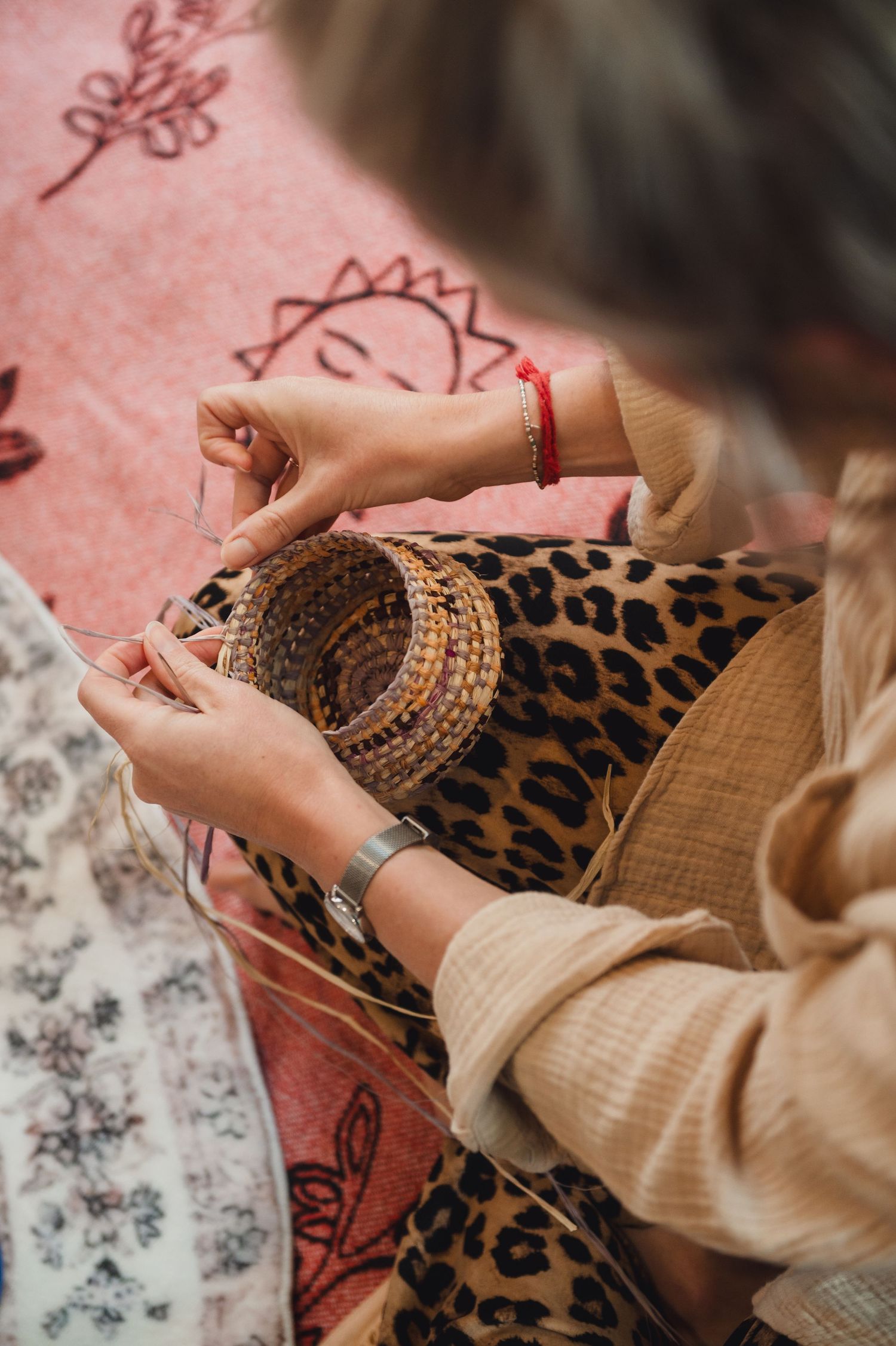 Woman weaving a small wicker basket by hand, wearing a leopard print top and red bracelet, seated on a pink patterned blanket.