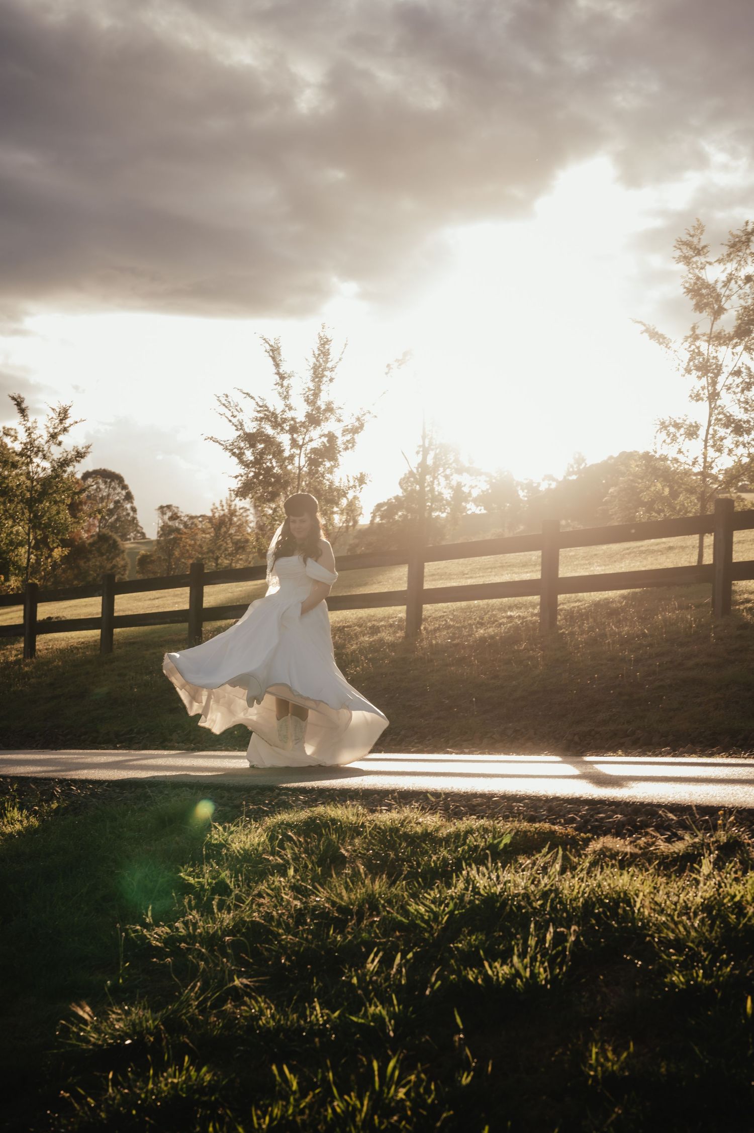 Bride in flowing white wedding dress twirls at golden sunset by a rustic wooden fence.