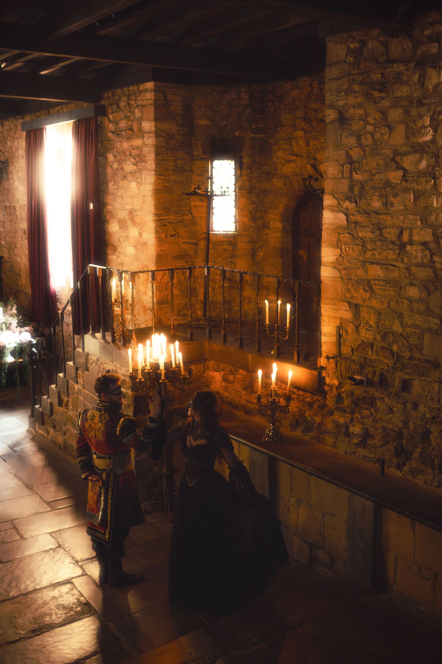 Medieval stone castle interior with candlelit altar, iron railings, and figures in period costume during a ceremonial gathering.
