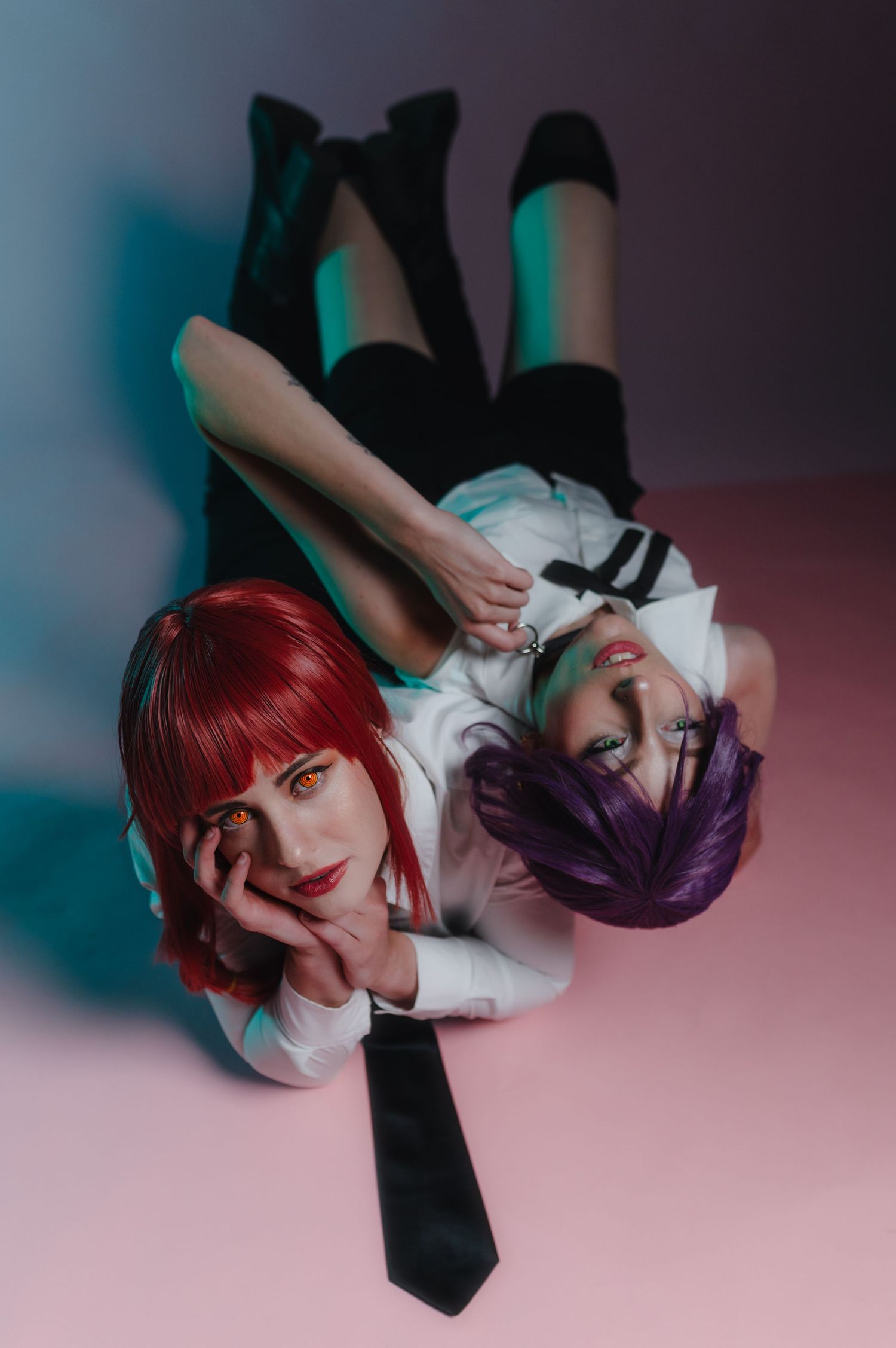 Two cosplay models with red and purple wigs in school uniforms lying on a pink floor under colorful studio lighting.