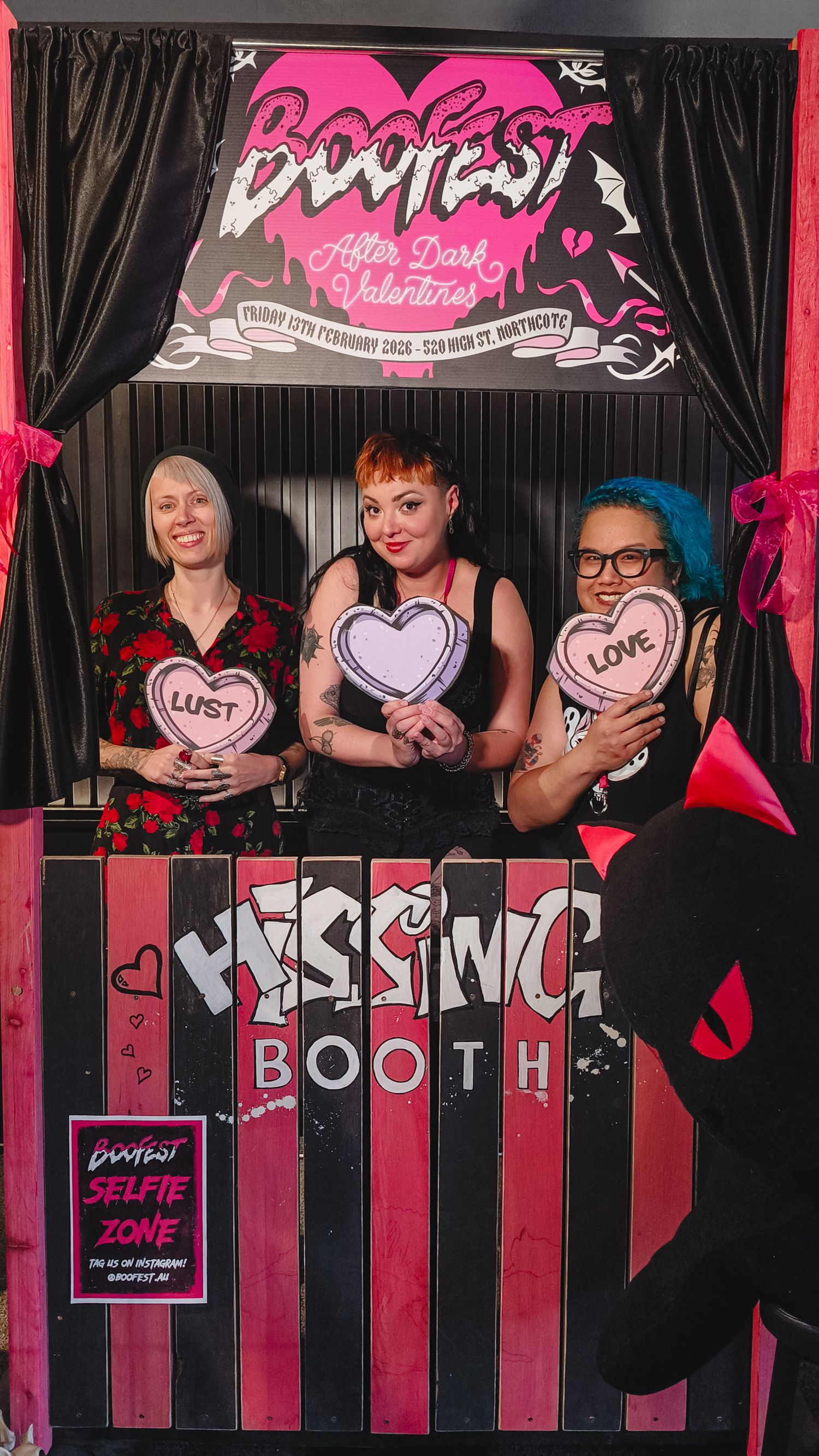 Three women pose at a Boofest After Dark Valentines kissing booth, holding heart signs reading Lust and Love.