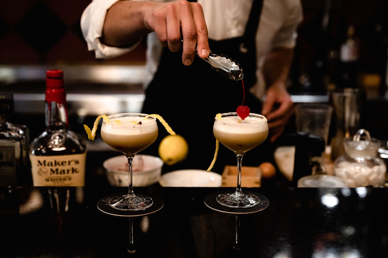 Bartender pouring a cherry garnish on creamy whiskey sour cocktails with Maker's Mark bourbon at a bar.