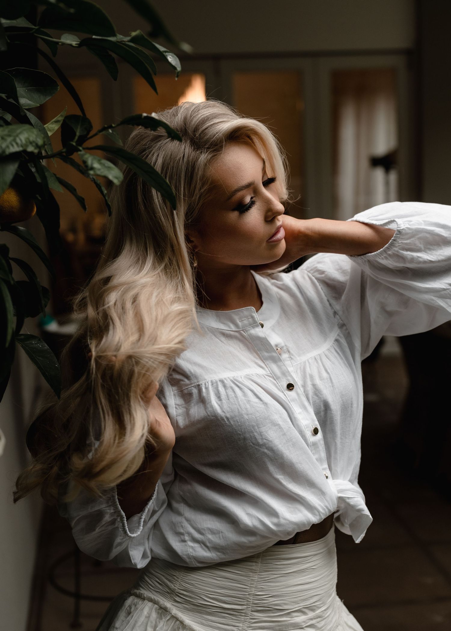 Blonde woman in white linen blouse poses gracefully beside tropical plant in elegant indoor setting.
