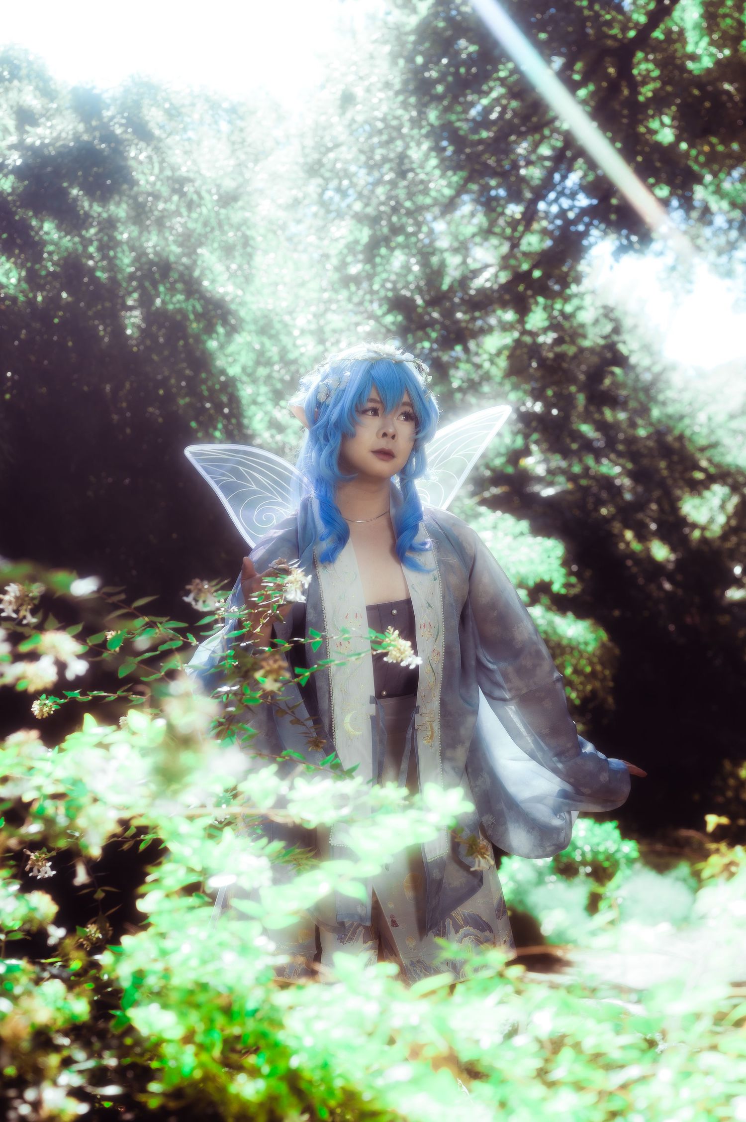 A cosplayer with blue hair and fairy wings poses in a sunlit forest wearing a silver and white ethereal costume.