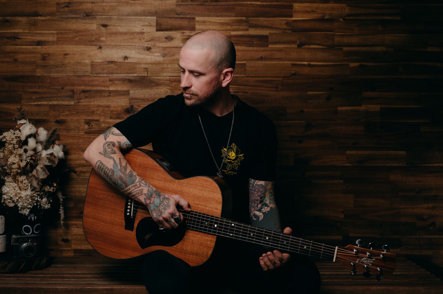 A musician plays acoustic guitar against a wooden wall backdrop.
