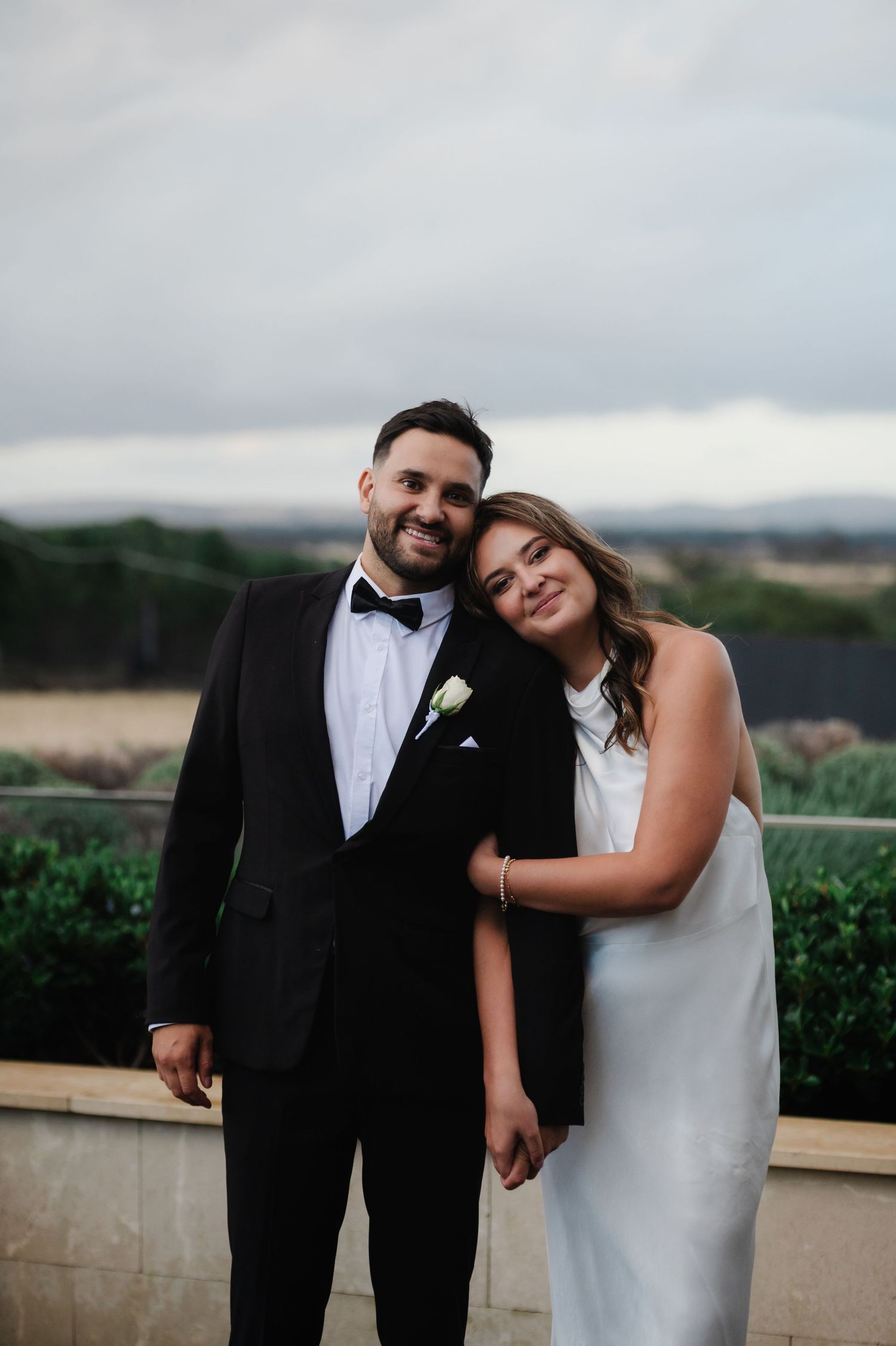 Couple in formal attire share intimate moment on winery terrace overlooking valley.