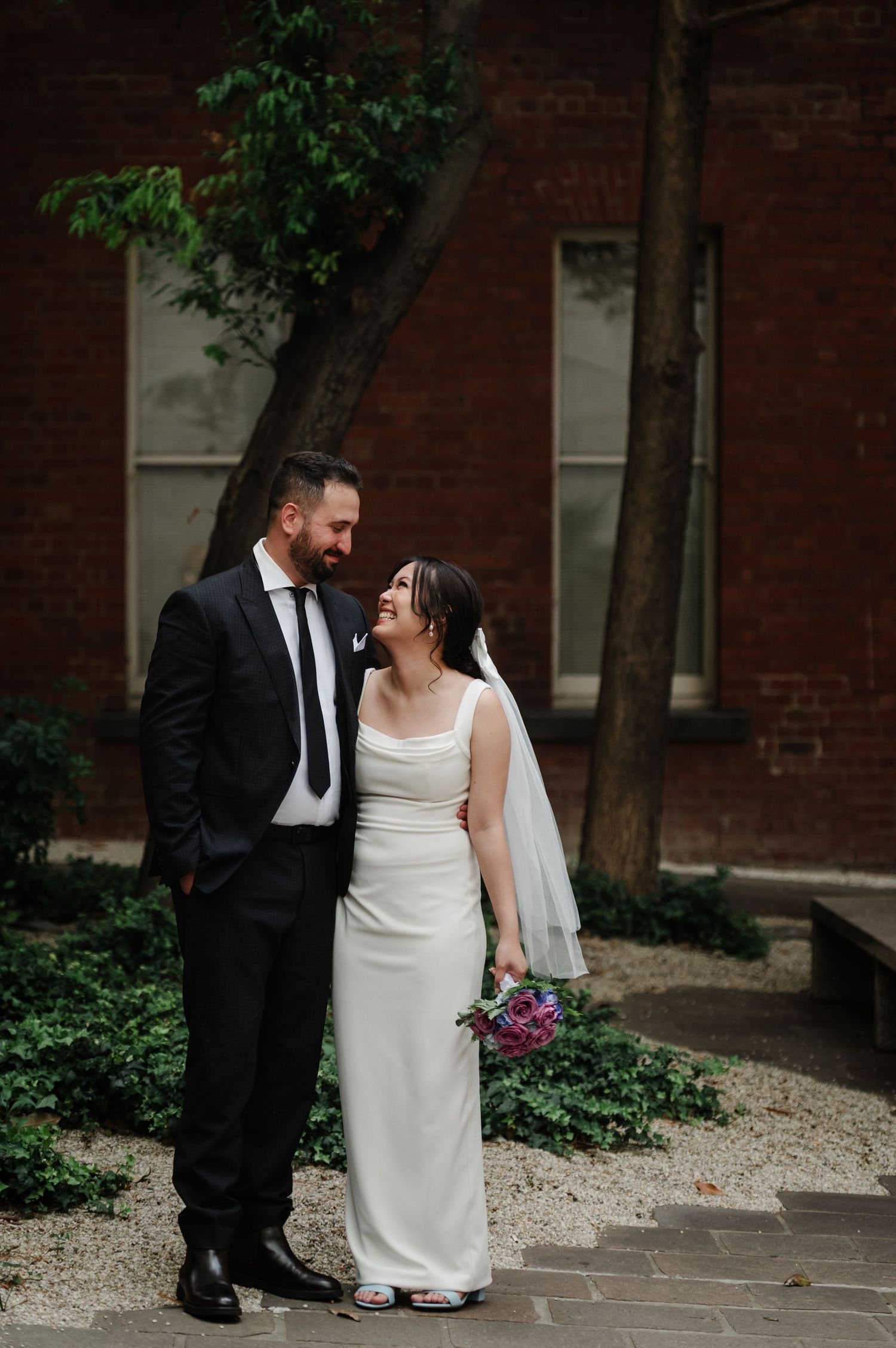 Newlywed couple shares an intimate moment against a brick wall backdrop with greenery.
