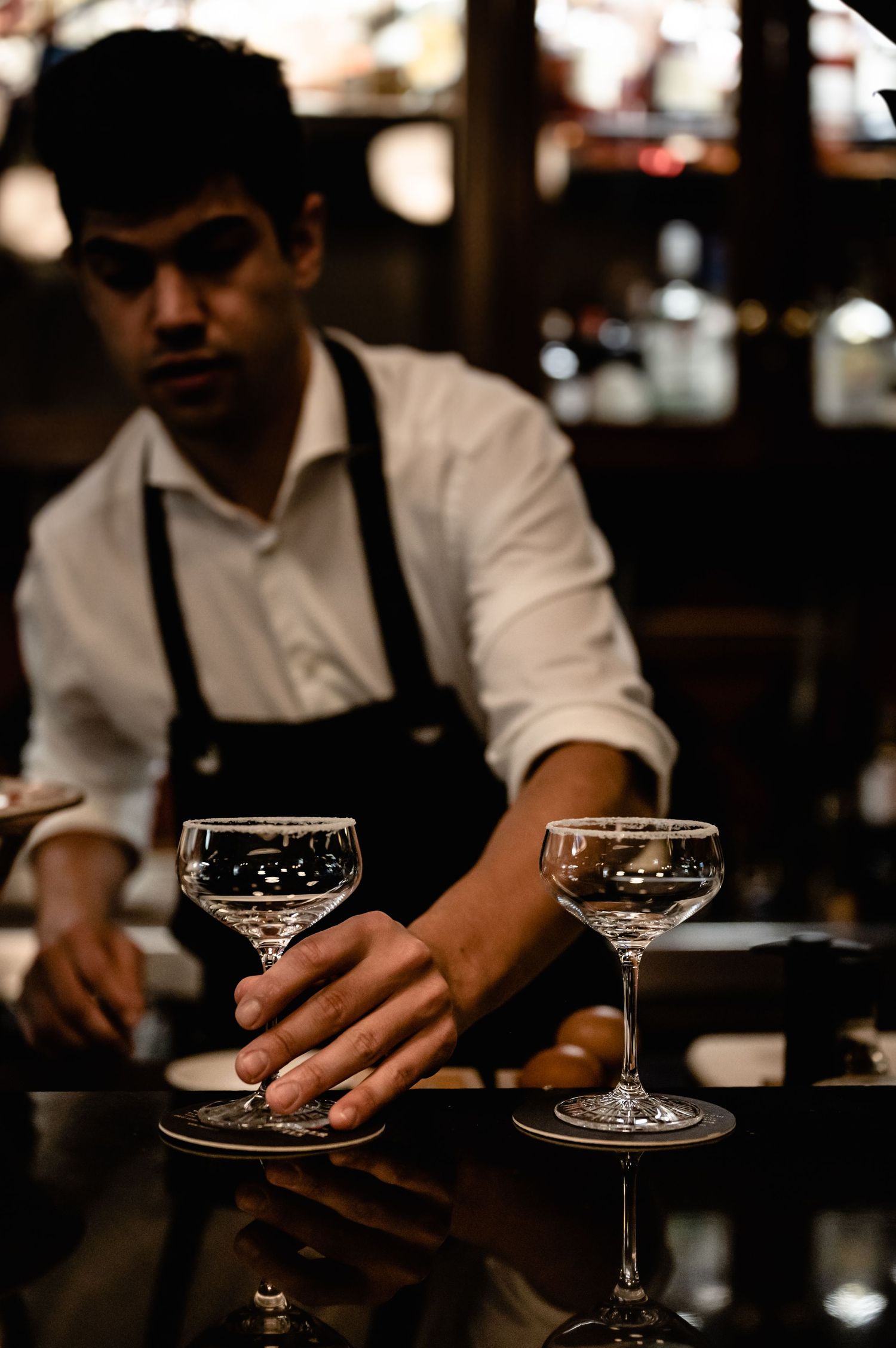 Bartender in white shirt and black apron placing crystal coupe glasses on a dark bar counter.