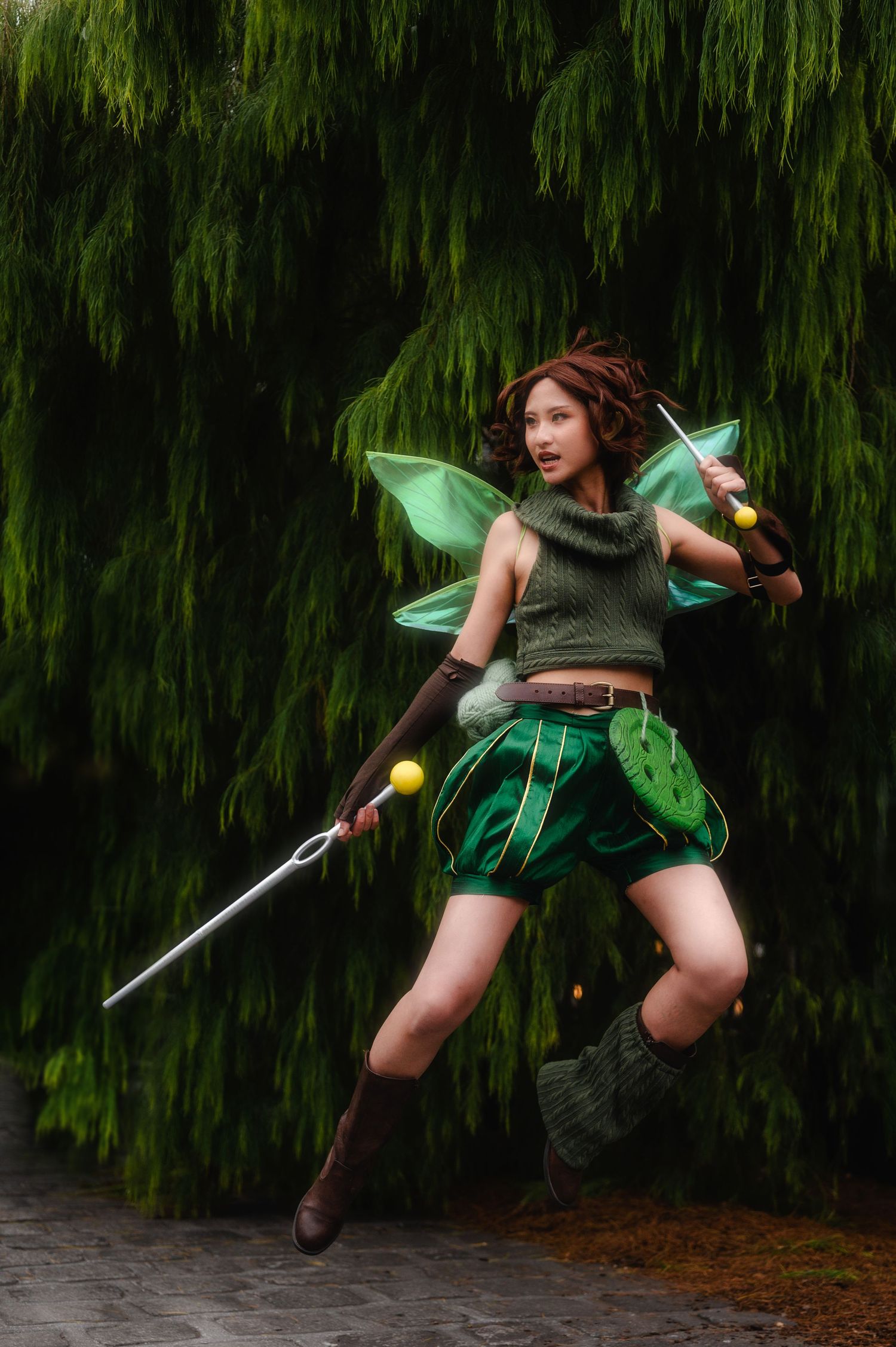 Woman cosplaying as a green fairy warrior, wielding a staff and sword, posing mid-jump before lush green foliage.