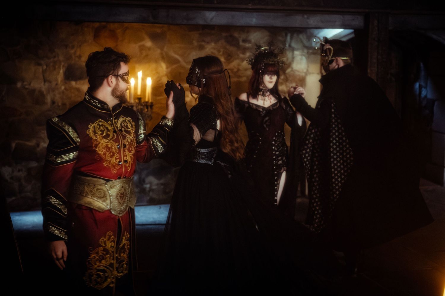 Four people in elaborate medieval fantasy costumes gather in a candlelit stone room during a dramatic role-playing event.