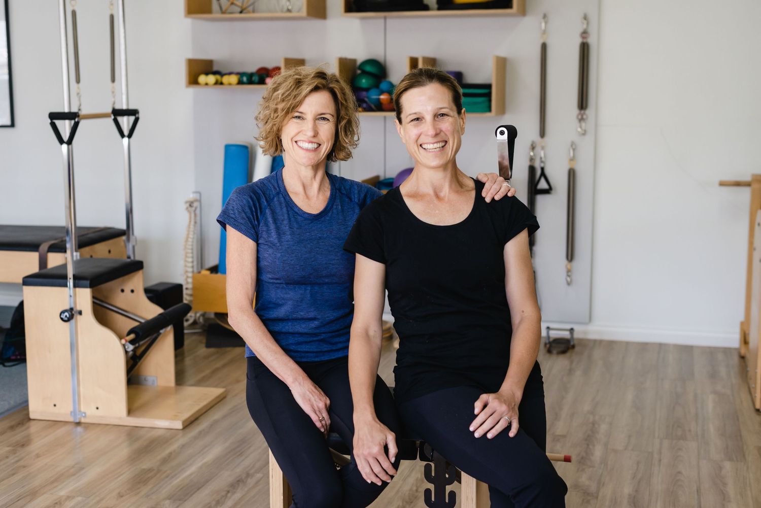 Two people pose together in a bright indoor studio space with fitness equipment visible in the background.