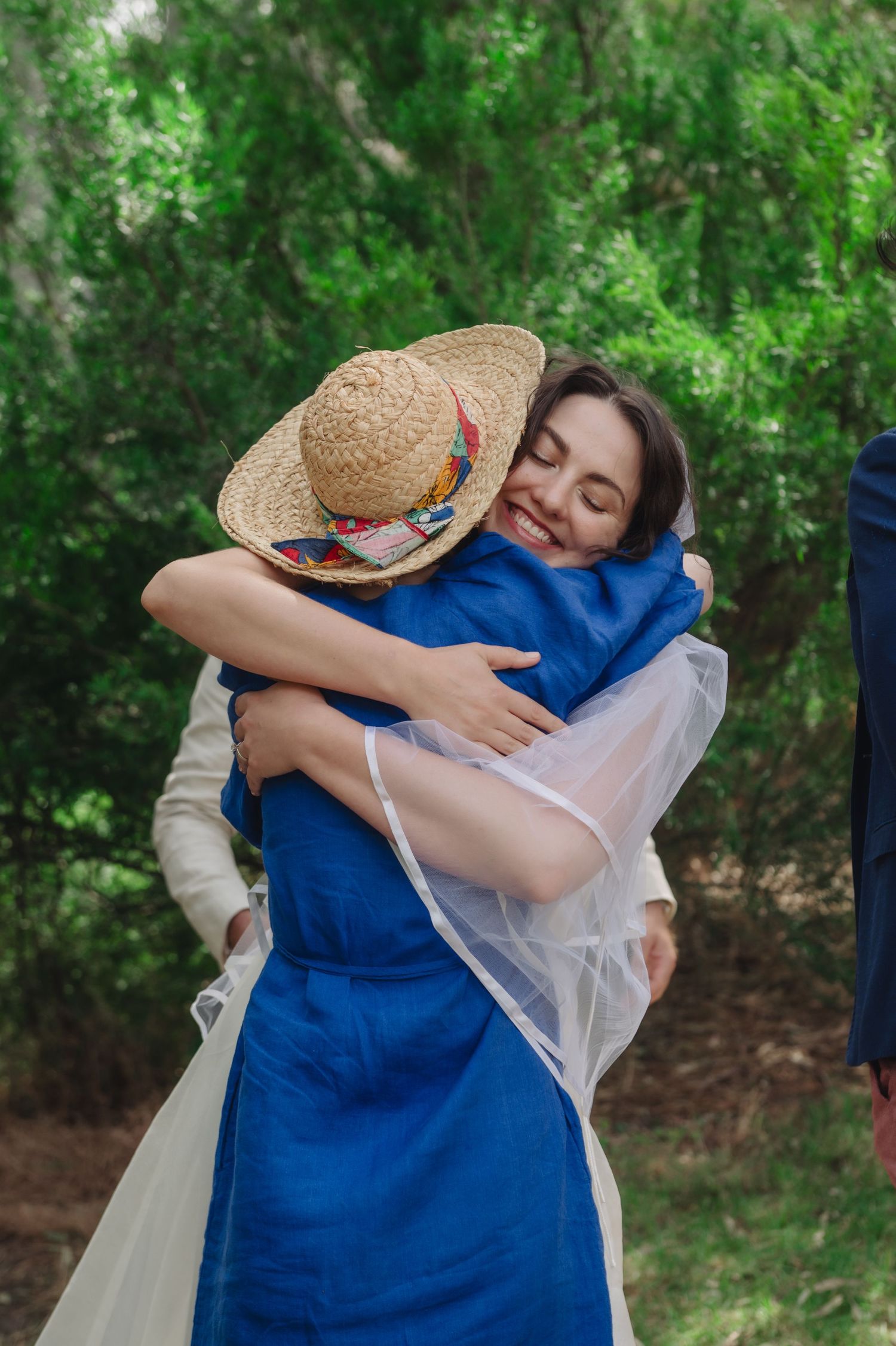 A smiling bride in a veil hugs a woman in a blue dress and straw hat outdoors amid lush green trees.
