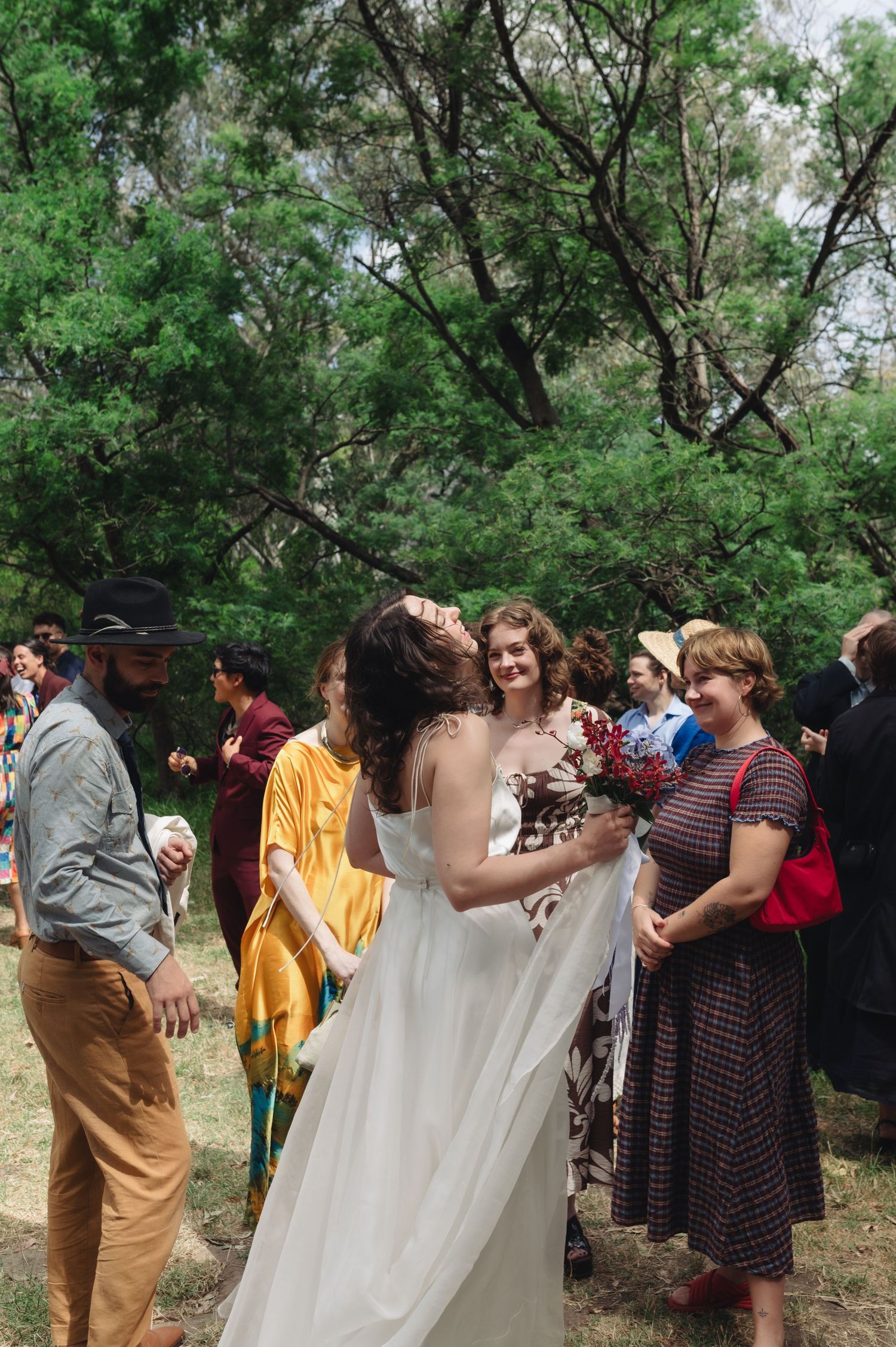 Bride in white gown laughs with guests during outdoor wedding reception surrounded by lush green trees.