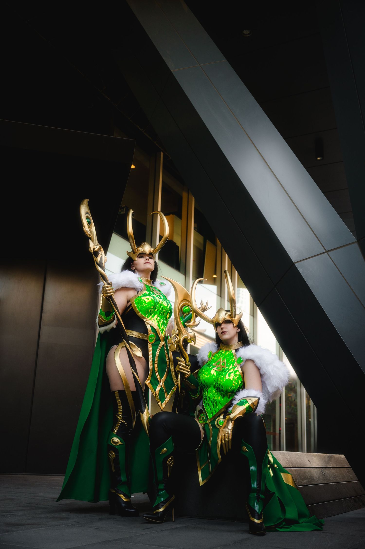 Two cosplayers in green and gold Loki costumes with horned helmets pose dramatically at a modern building.
