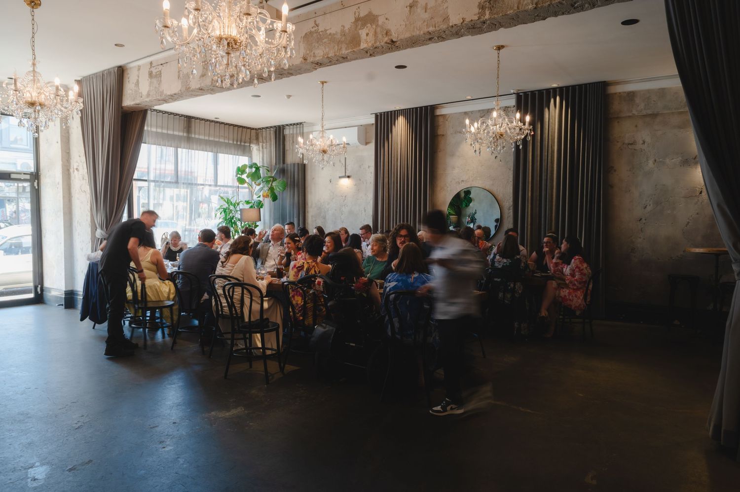 Elegant event space with crystal chandeliers and exposed brick walls features guests seated at round tables.