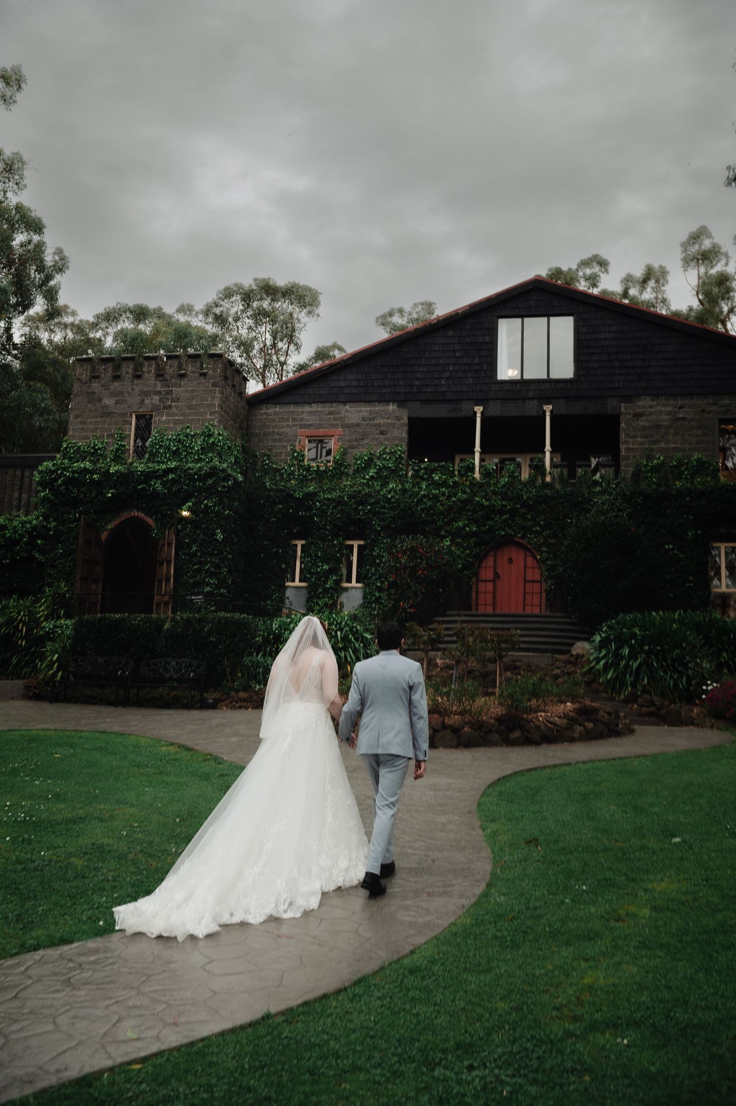Couple walks along curved pathway towards rustic barn venue on overcast day.