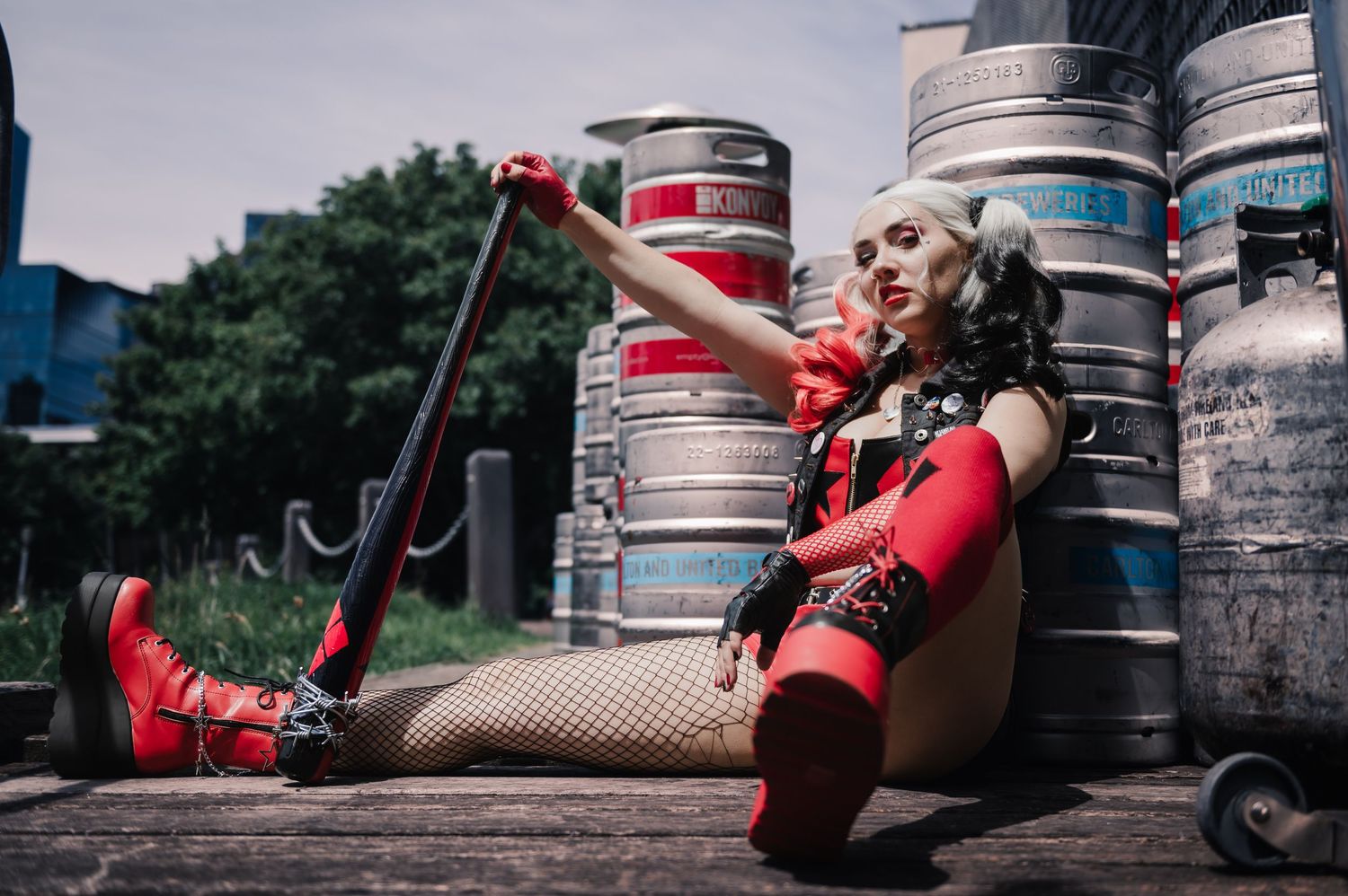 Urban cosplay photoshoot featuring red and black outfit against industrial metal kegs.