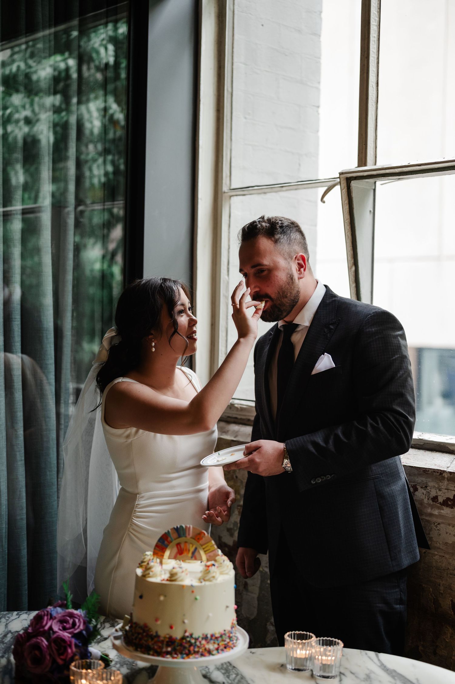 Newlyweds laughing while feeding each other wedding cake.