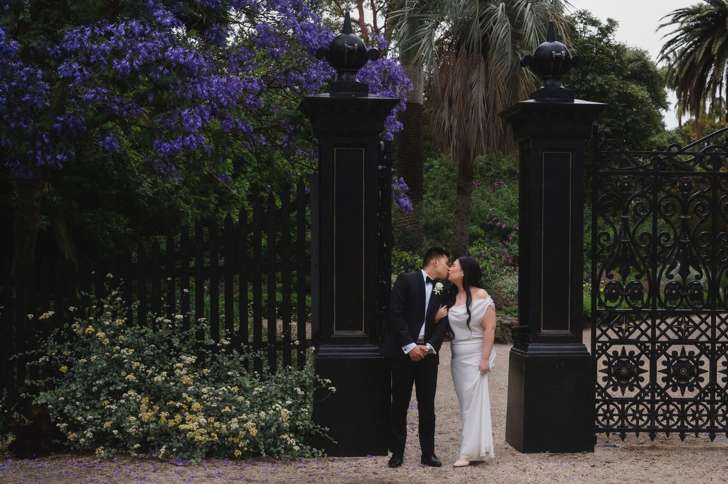 A couple poses for wedding photos in a lush garden setting with blooming purple jacaranda trees and palm trees.