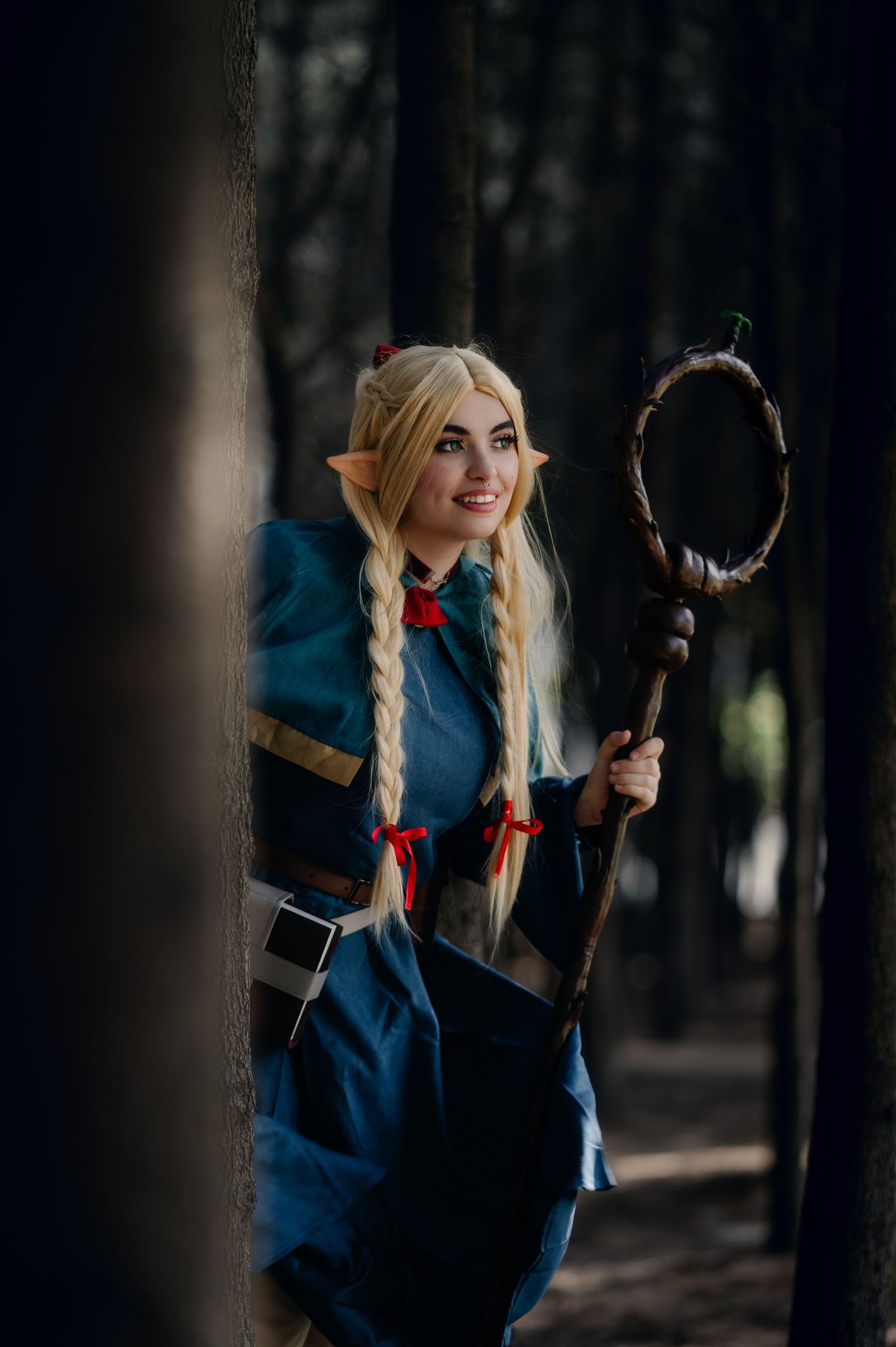 Cosplayer in blue dress with blonde braids poses with staff in moody forest setting.