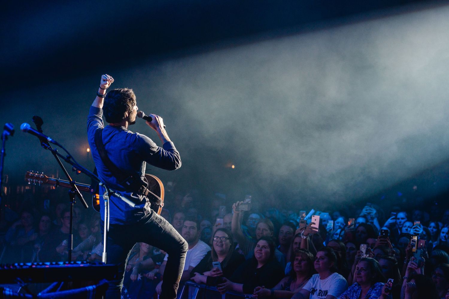 A rock musician performs on stage with fist raised, facing an enthusiastic crowd under dramatic spotlights and smoke.