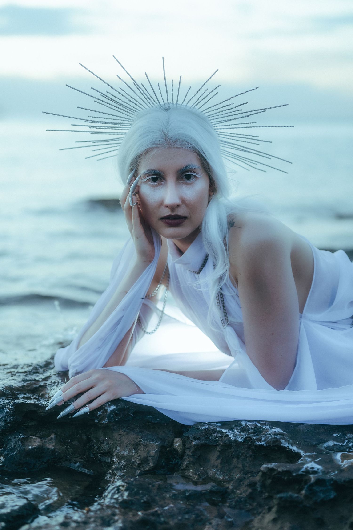 Woman in white dress and spiked halo crown poses on ocean rocks at dusk in ethereal fantasy portrait.
