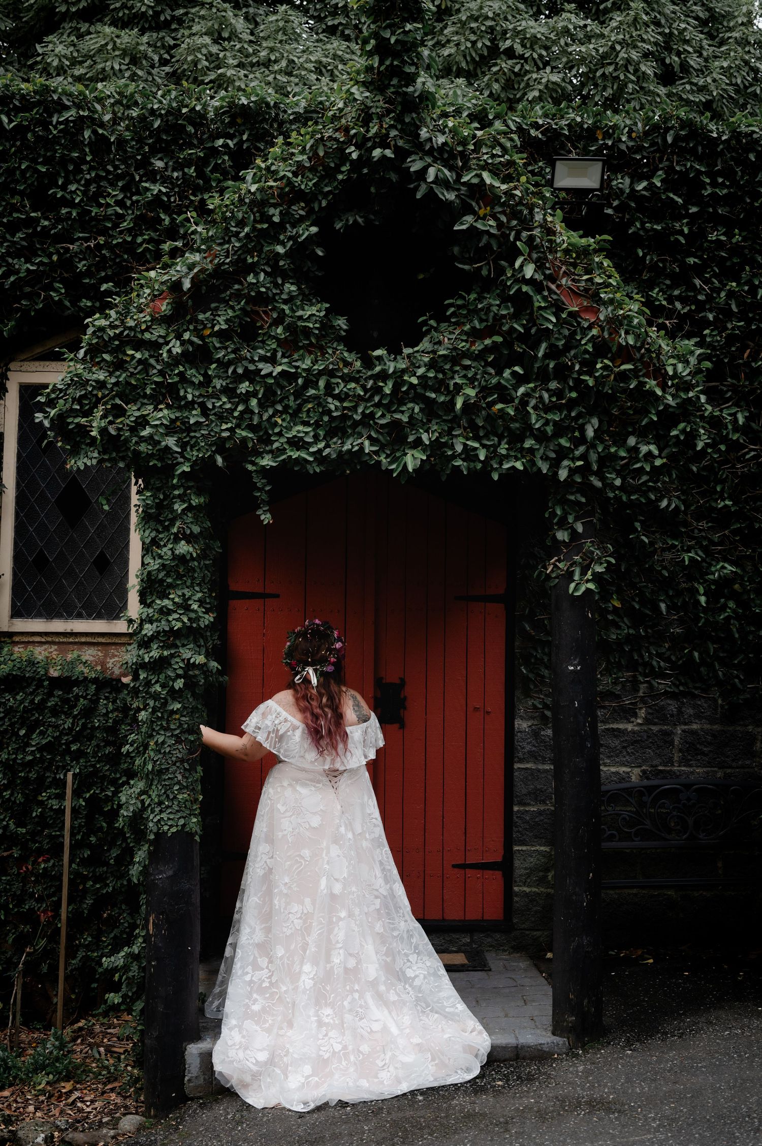 Bride in lace gown poses by red door surrounded by lush green ivy.