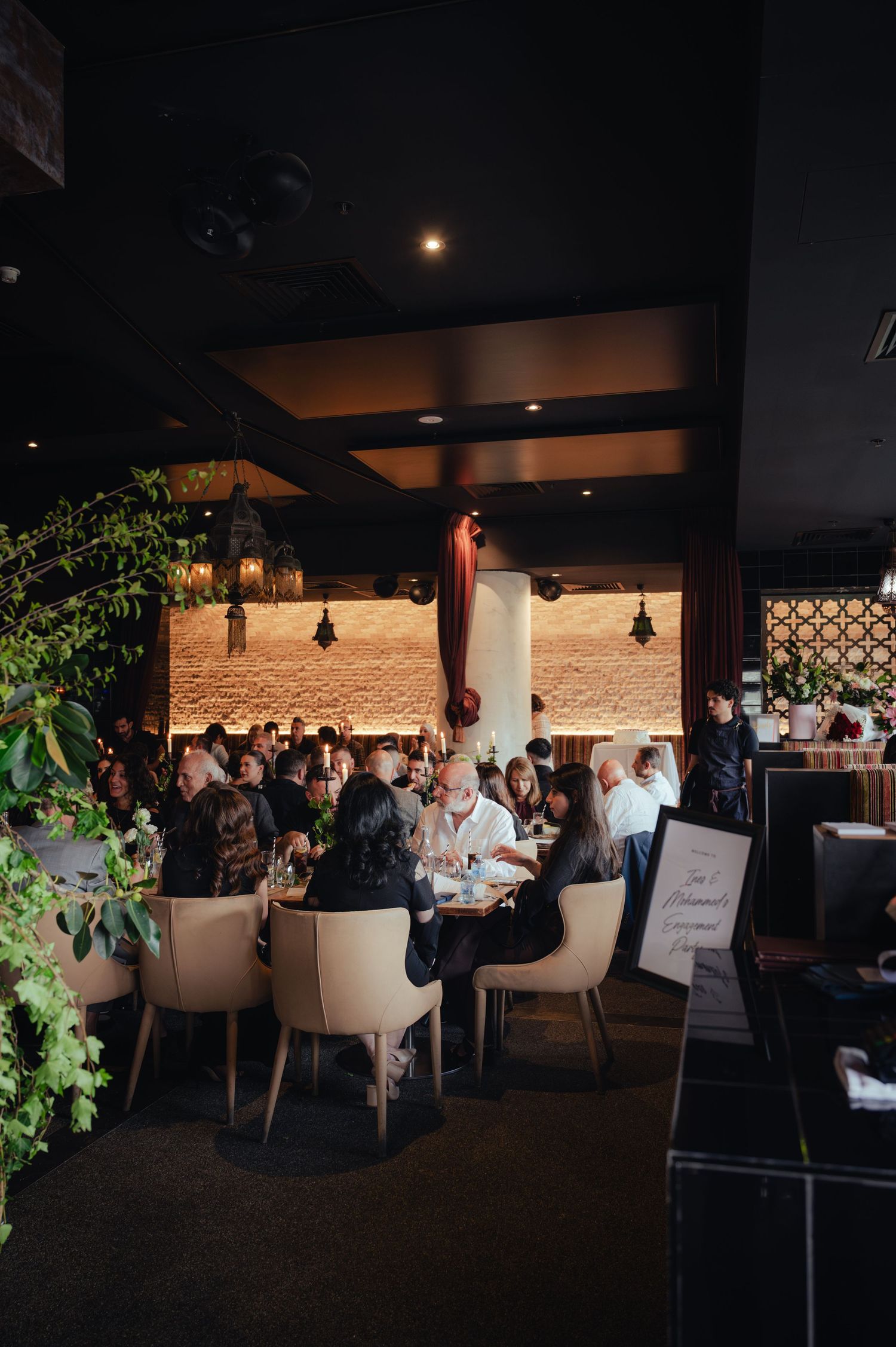 Elegant restaurant dining room with guests seated at round tables, warm ambient lighting, and lush greenery accents.