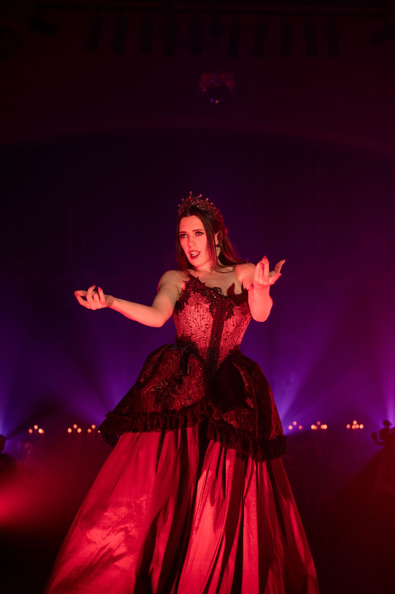 Woman in red and black ball gown with dark crown poses dramatically on stage under purple and red lights.
