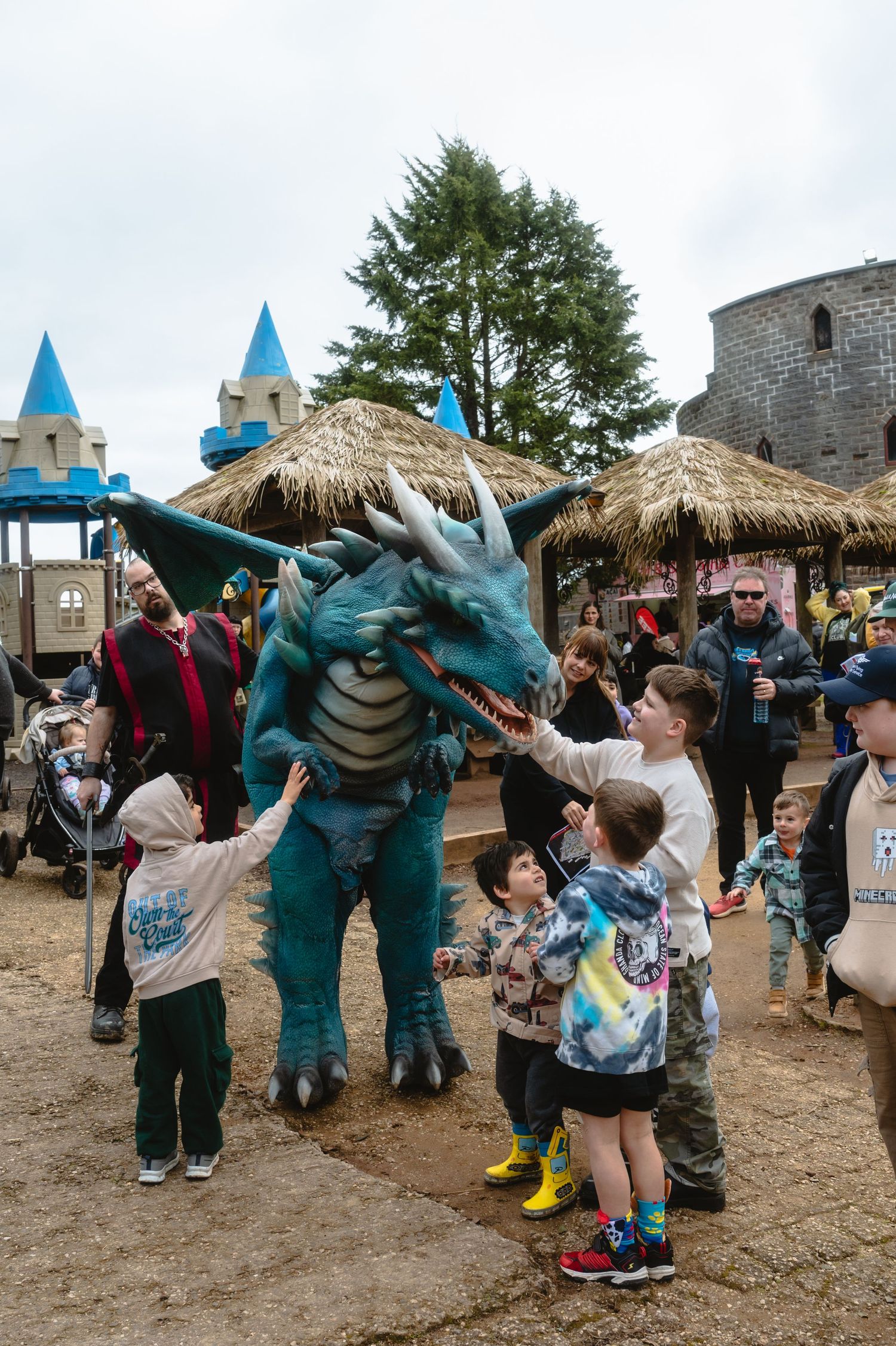 Children interact with a teal dragon costume character at a medieval-themed amusement park with castle decorations.