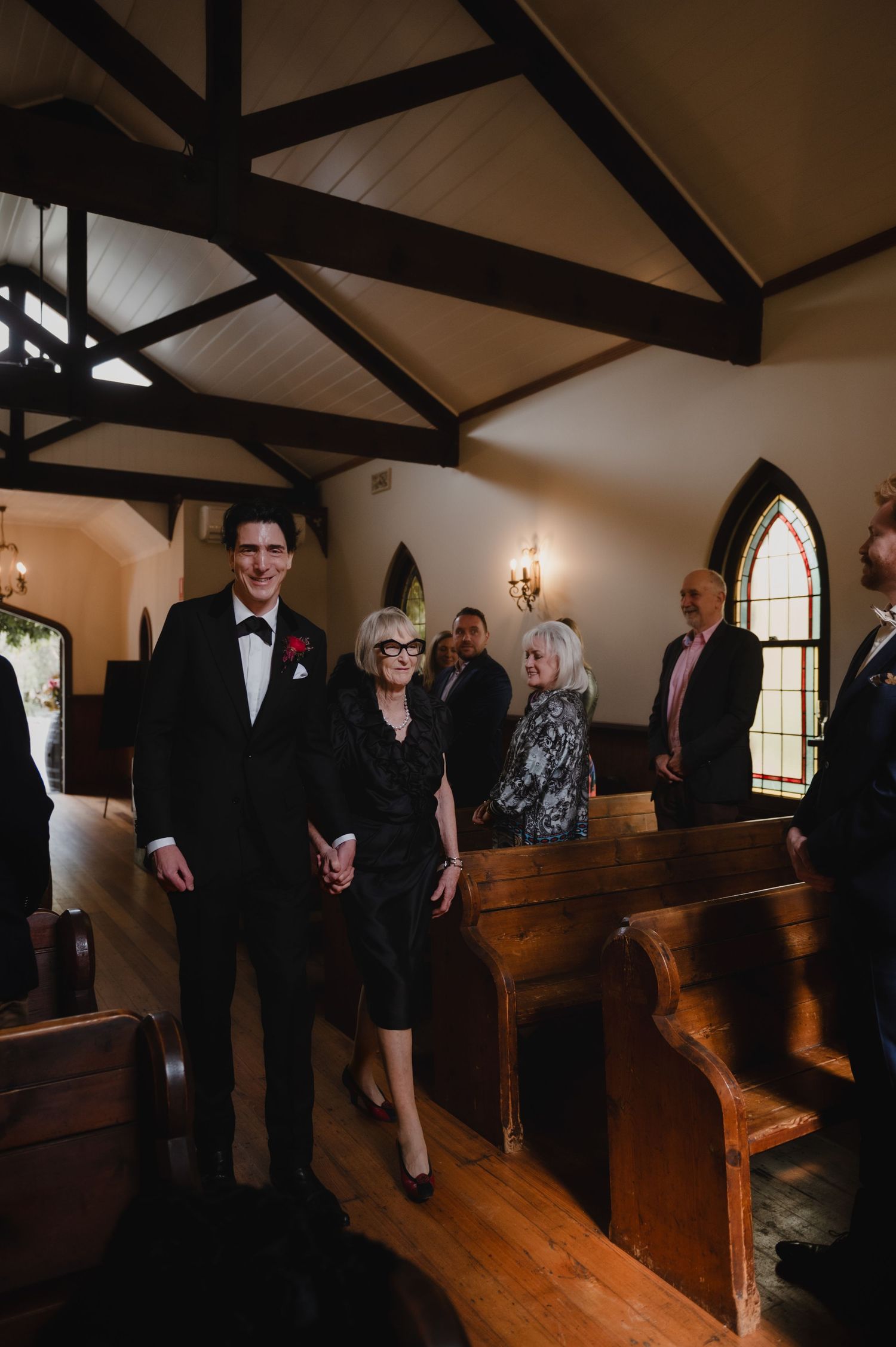 Wedding guests gather in a rustic chapel with wooden pews and exposed ceiling beams.