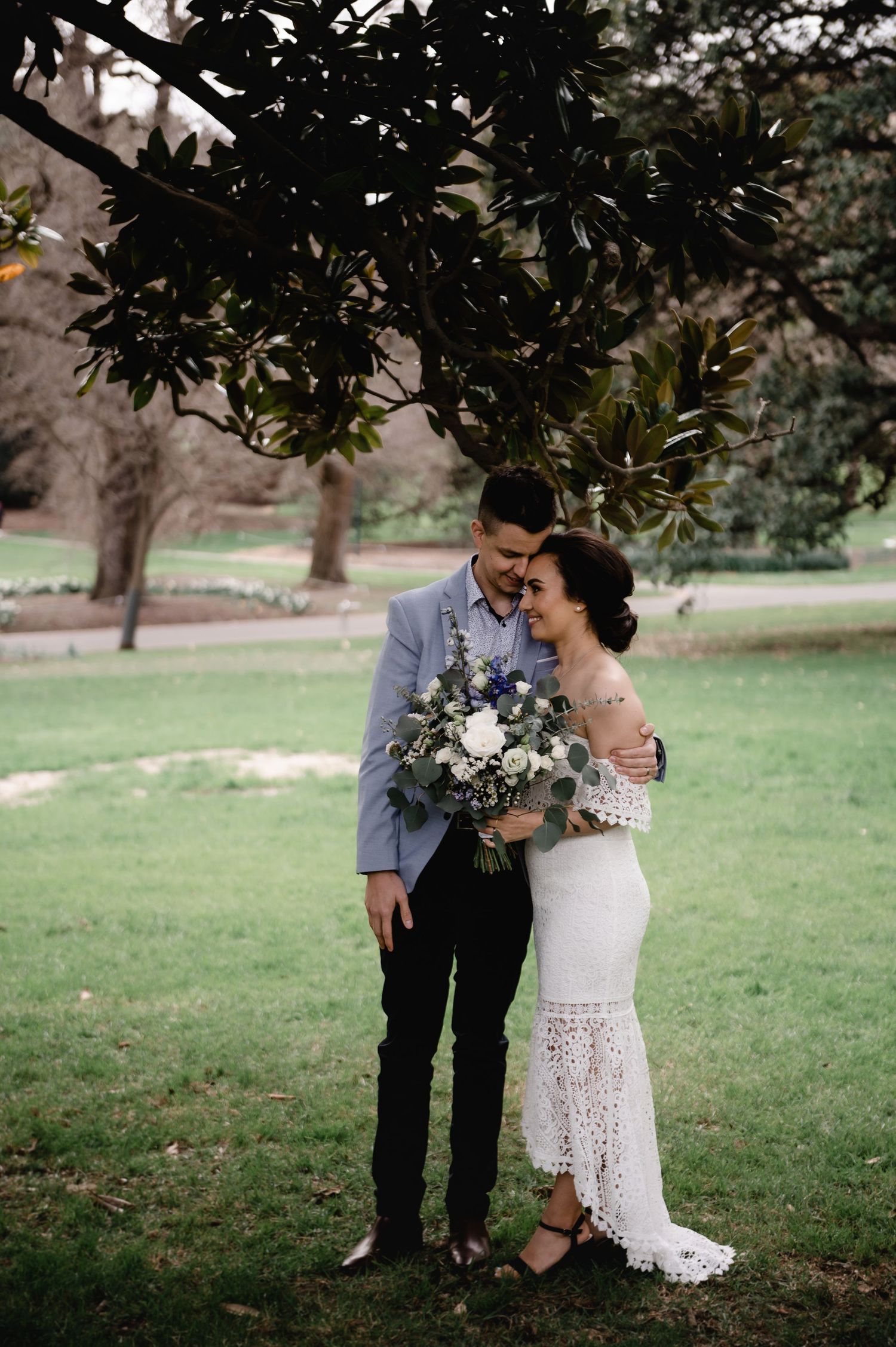 A romantic wedding couple embraces under tree branches in a garden setting with a beautiful floral bouquet.