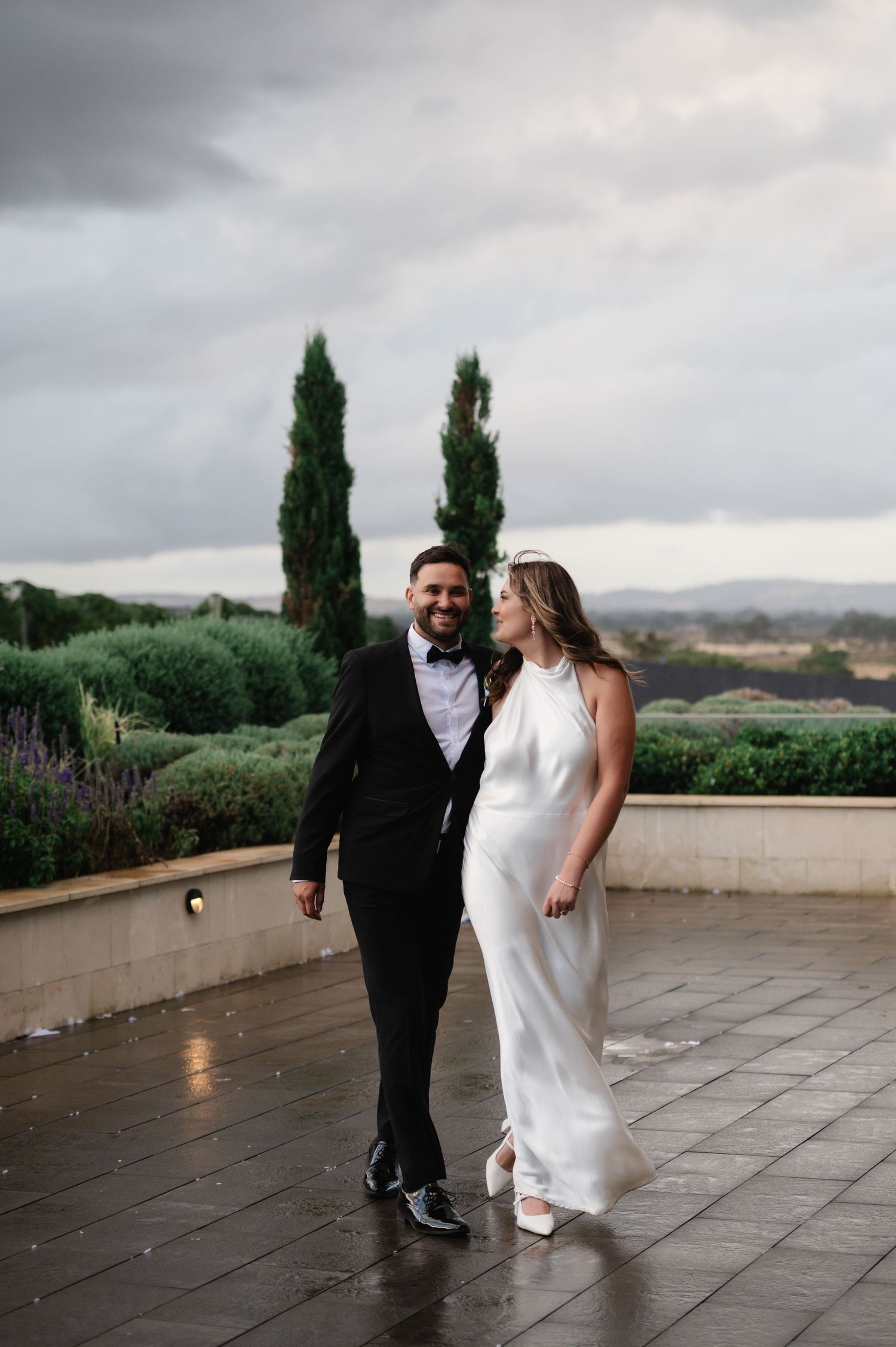Newlyweds pose playfully on stone terrace with cypress trees and rolling hills in background.