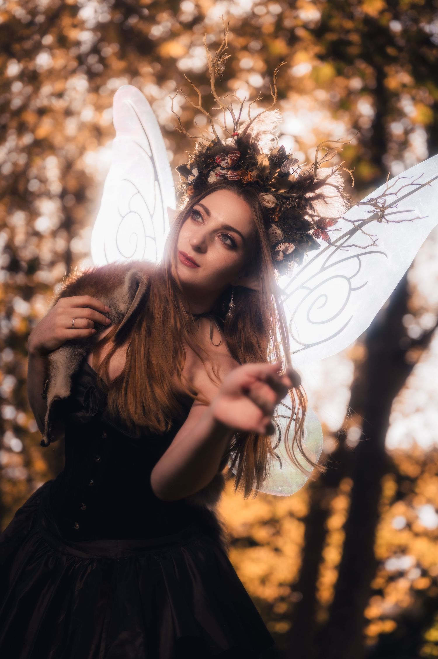 A woman in a dark fairy costume with white wings poses in an autumn forest wearing a floral crown.