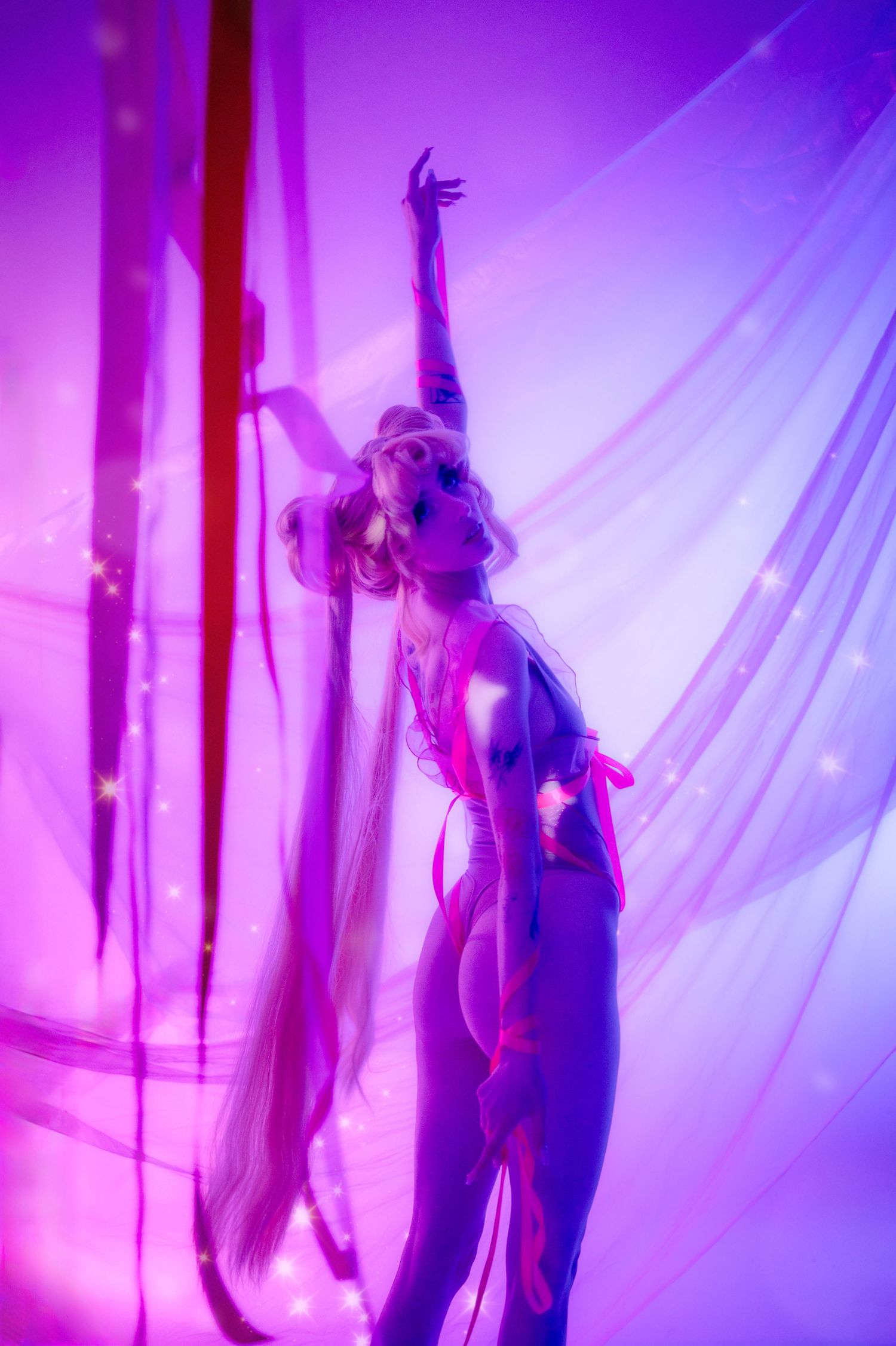 A performer poses dramatically under purple and pink lights with flowing sheer fabric and twinkling stars.