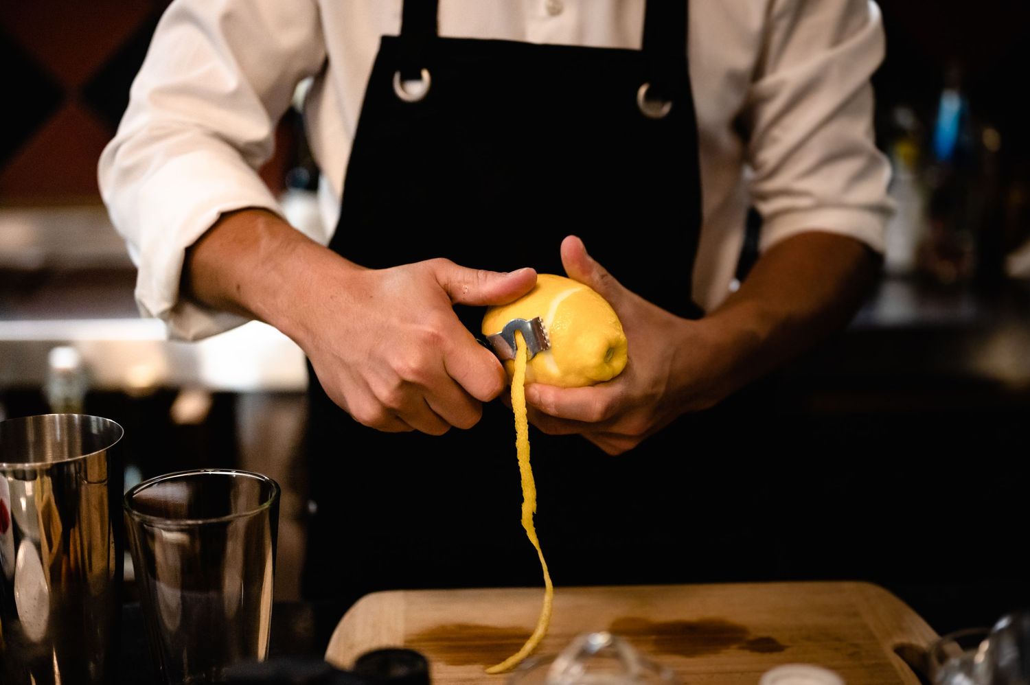 Chef in black apron peeling a fresh lemon with a peeler over a wooden cutting board in a professional kitchen.