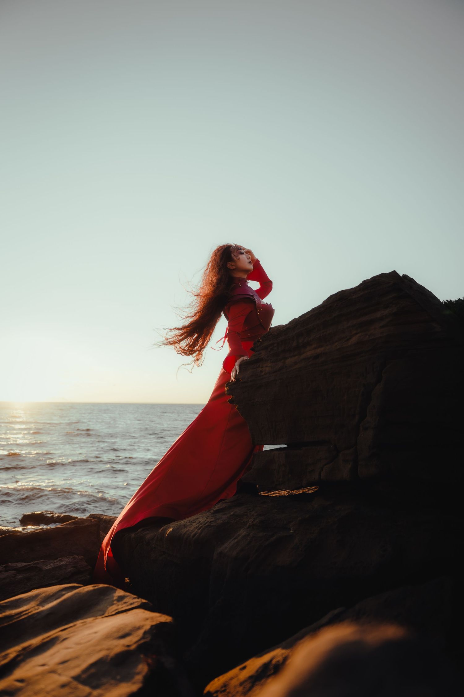 Figure in flowing red dress silhouetted against ocean sunset on rocky cliff.