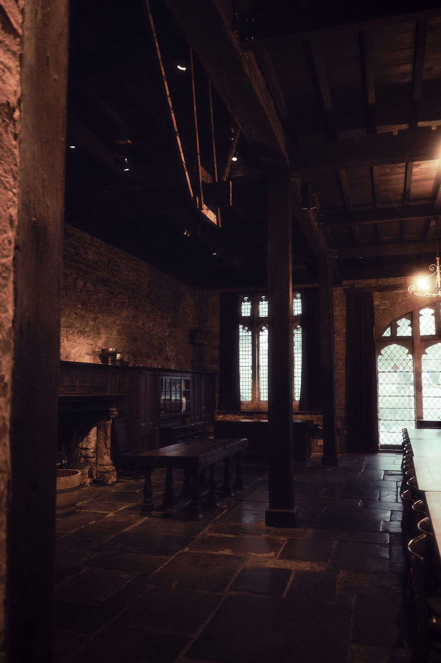 Dark medieval hall with stone fireplace, Gothic windows, wooden beamed ceiling, and long dining table on slate floor.