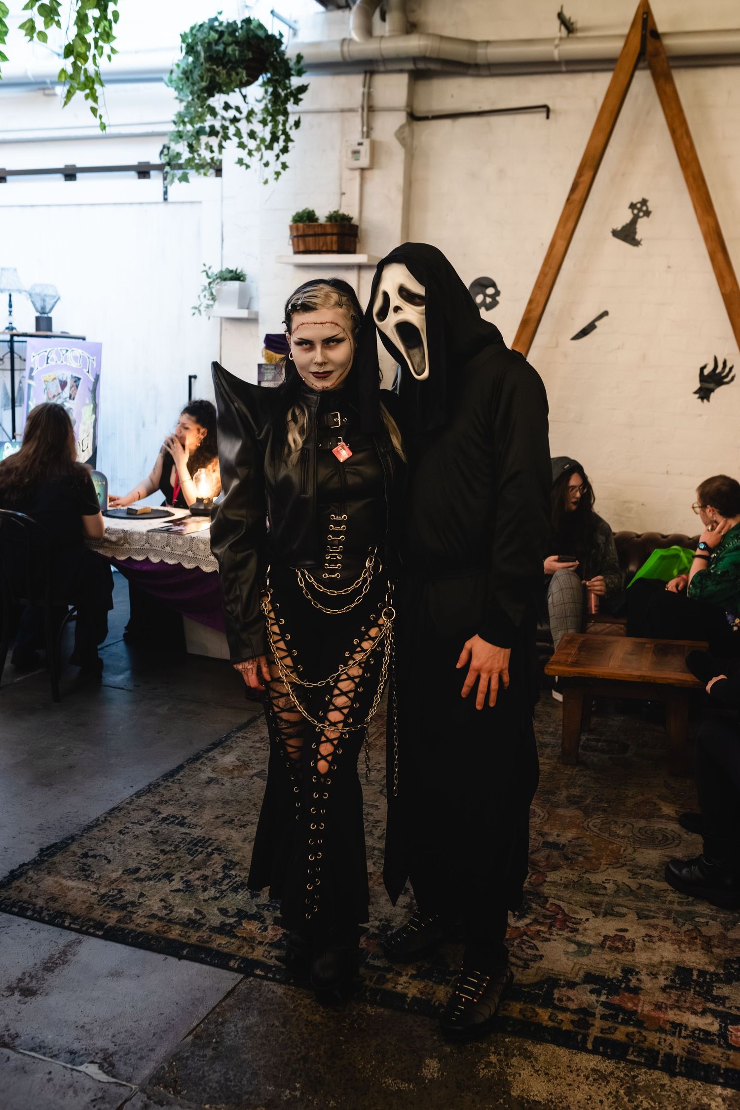 Two people in Halloween costumes, one as a dark witch and one as Ghostface from Scream, pose at an indoor party.
