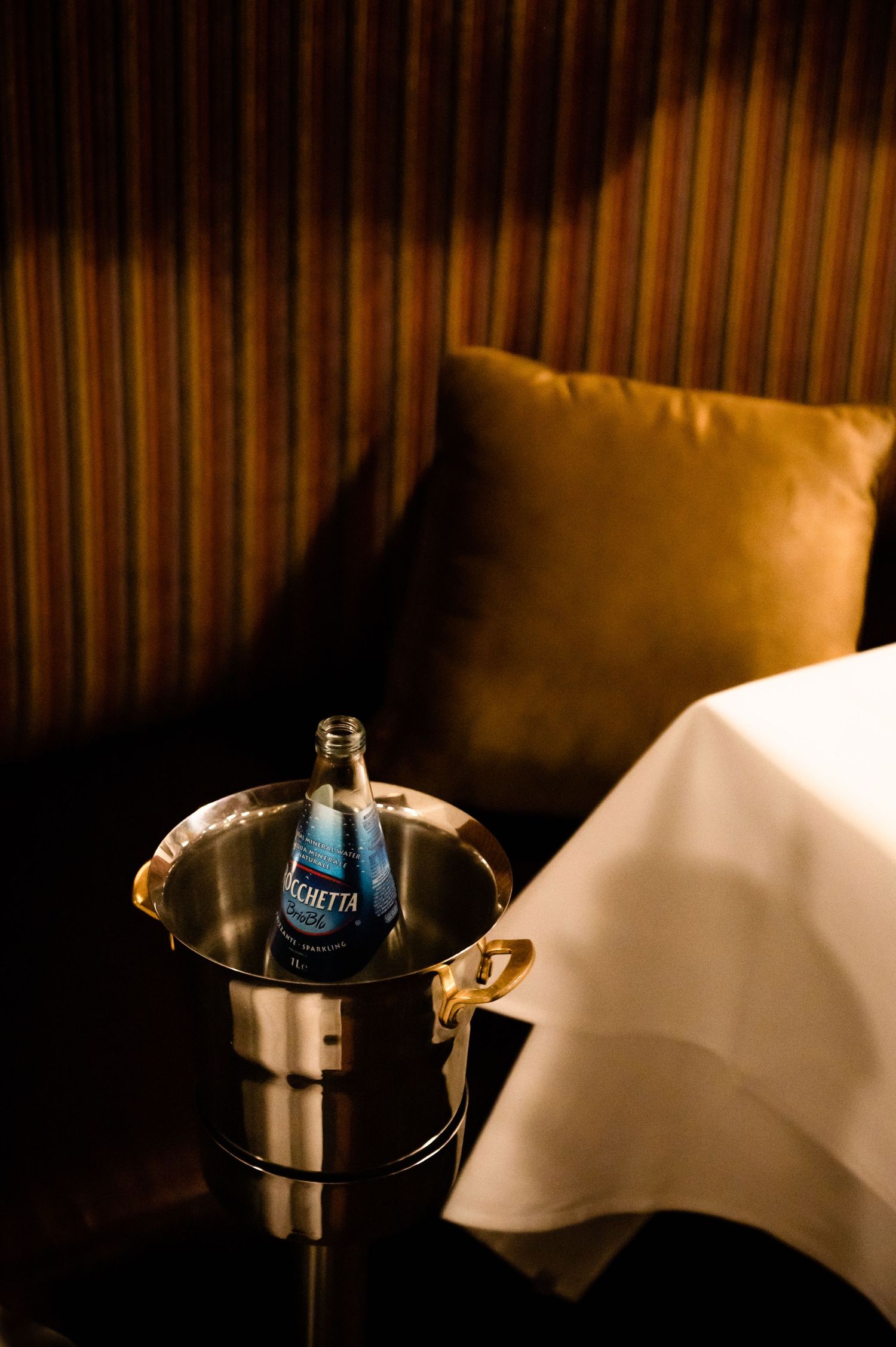 A bottle of Ferrarelle water chilling in a silver ice bucket beside a bed with white linens in a luxury hotel room.