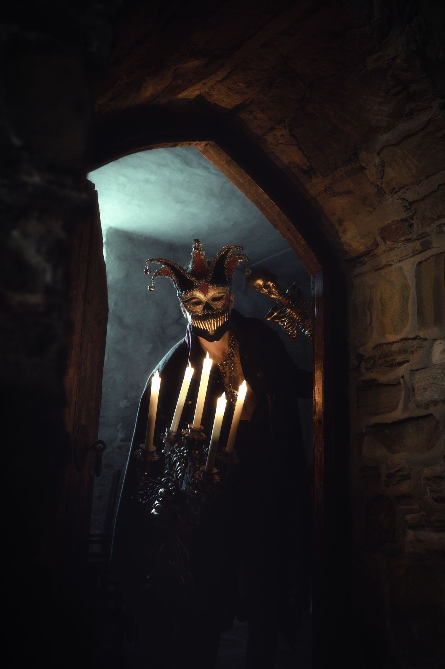 A creepy jester mask illuminated by candlelight in a dark stone archway.